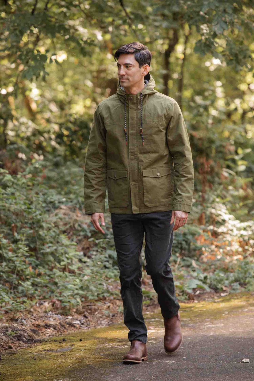 A man wearing the Bridge & Burn Marshall Jacket in olive waxed canvas, dark pants, and brown boots walks along a paved path bordered by lush greenery in a forest or park.