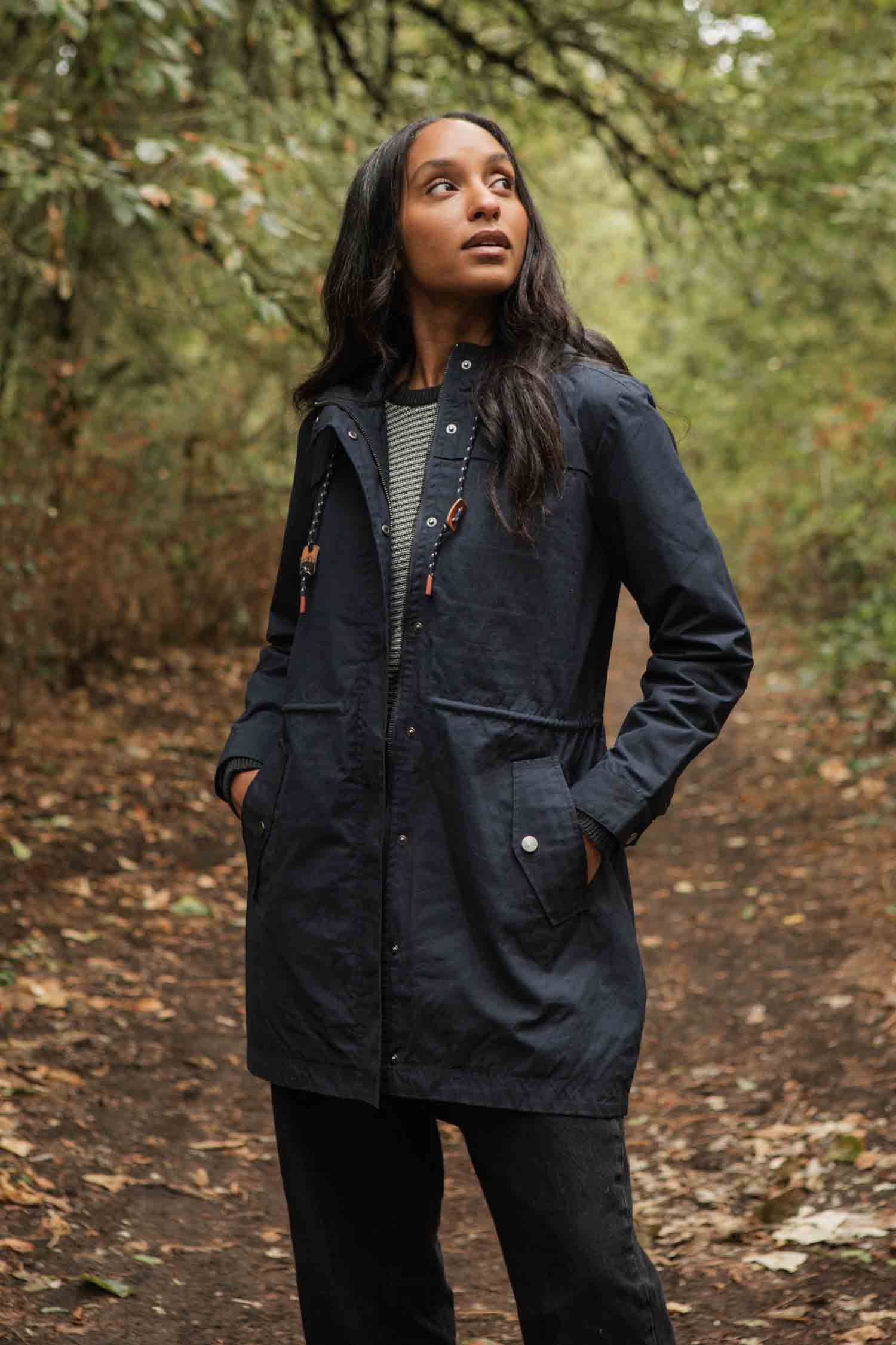 A woman with long dark hair stands on a forest path, hands in the pockets of her Bridge & Burn Briar Jacket in navy waxed canvas. She wears black pants and a striped shirt, surrounded by green trees and fallen leaves.