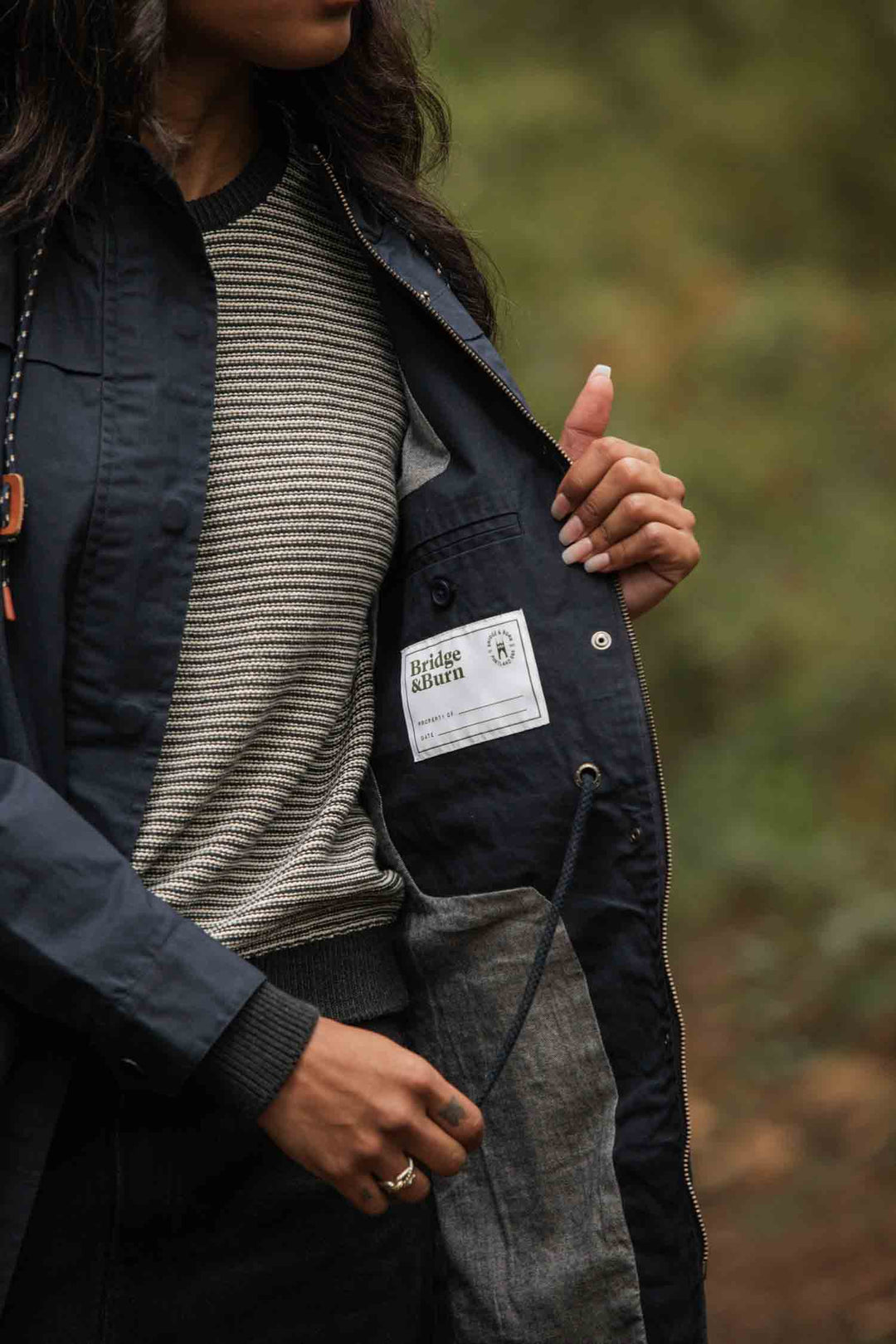 A person wearing a striped shirt and dark jacket stands outdoors, revealing the Bridge & Burn label inside the Briar Jacket / Navy Waxed Canvas—a rain-ready piece set against blurred greenery.