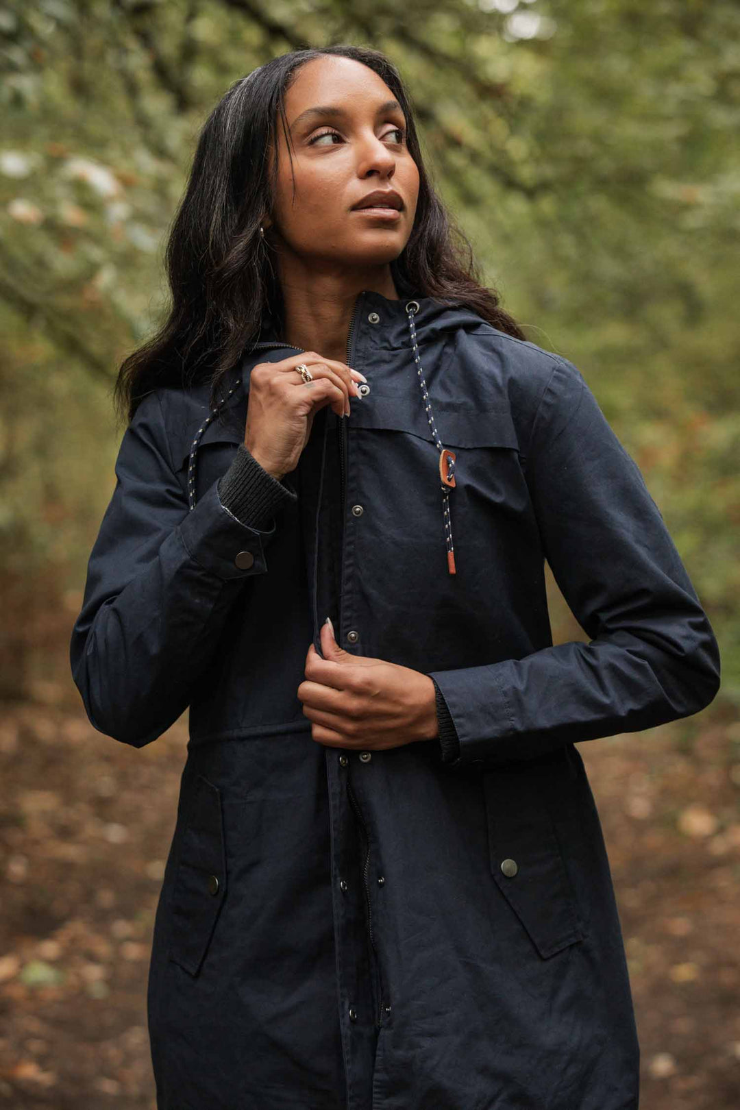 A woman wearing the Bridge & Burn Briar Jacket in navy waxed canvas stands outdoors on a forest path, holding the drawstrings and looking up to her left. The background features blurred green foliage.