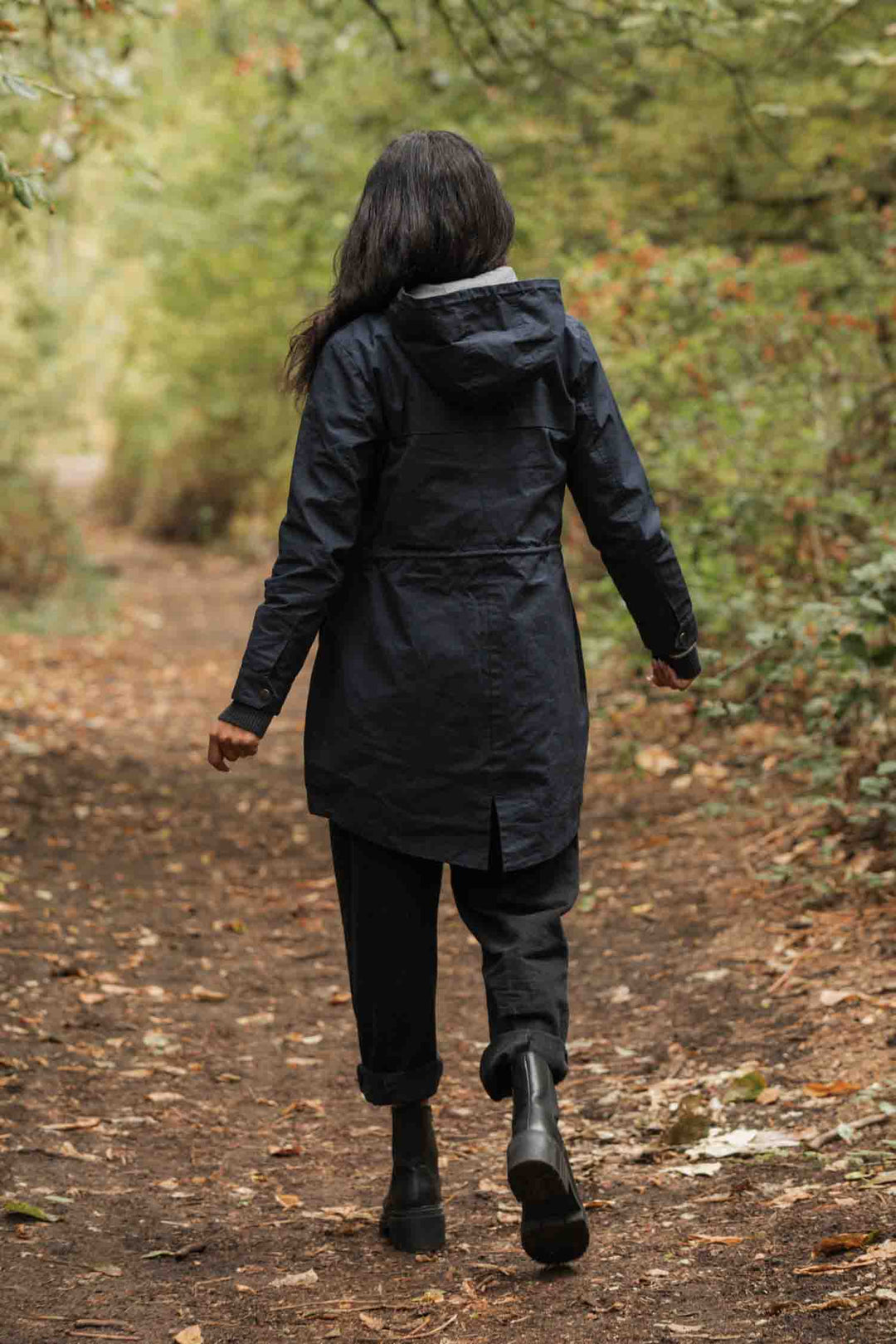 Wearing the Bridge & Burn Briar Jacket in navy waxed canvas, a person with long dark hair walks away on a leafy forest path surrounded by greenery.