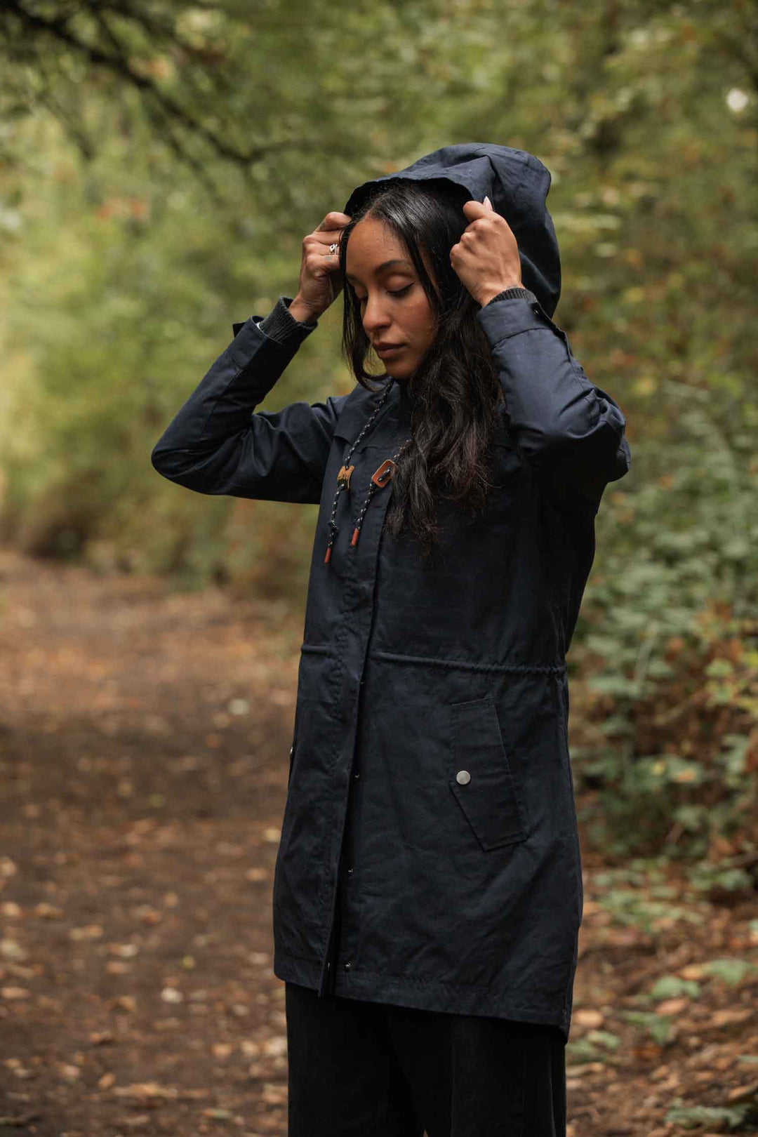 A woman stands on a forest path, adjusting the hood of her Bridge & Burn Briar Jacket in navy waxed canvas. Lush green foliage surrounds her, and fallen leaves cover the ground.