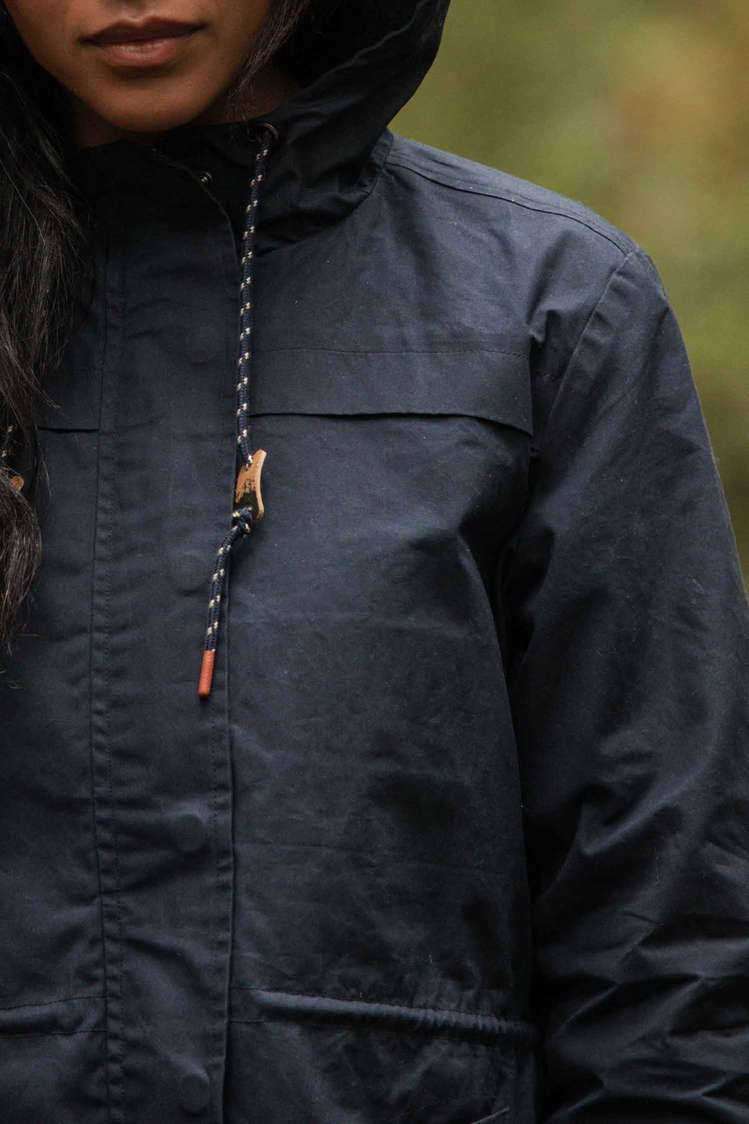 A person stands outdoors wearing the Bridge & Burn Briar Jacket in navy waxed canvas, its hood and drawstrings visible. The image is cropped to show just the lower half of their face and upper body against a blurred natural background.