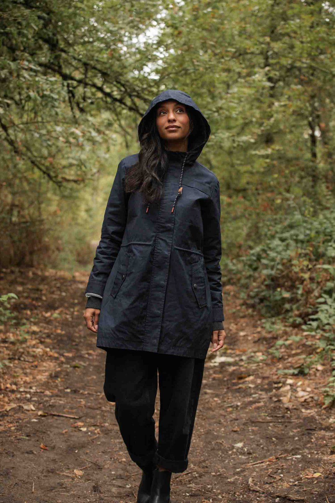 Wearing the Bridge & Burn Briar Jacket in navy waxed canvas, a person in black pants walks along a forest path lined with green trees and fallen leaves.
