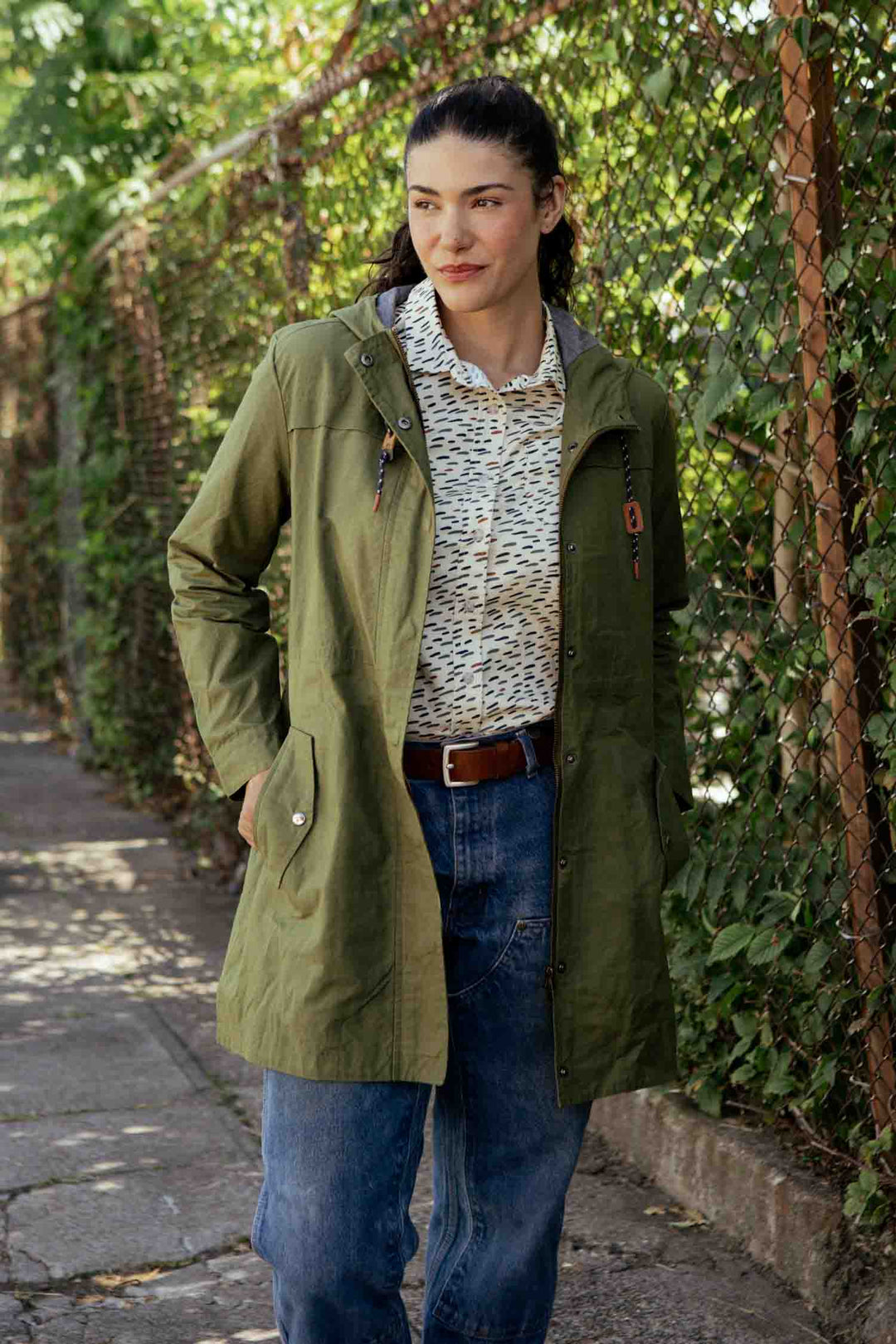 A woman stands outdoors by a chain-link fence covered in greenery, wearing the Bridge & Burn Briar Jacket in Olive Waxed Canvas over a patterned shirt and blue jeans, with her hands in her jacket pockets.
