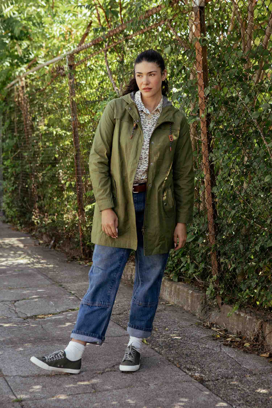 A woman stands on a sidewalk by a greenery-covered chain-link fence, wearing the Bridge & Burn Briar Jacket in Olive Waxed Canvas, along with rolled-up blue jeans, a patterned shirt, and gray sneakers.