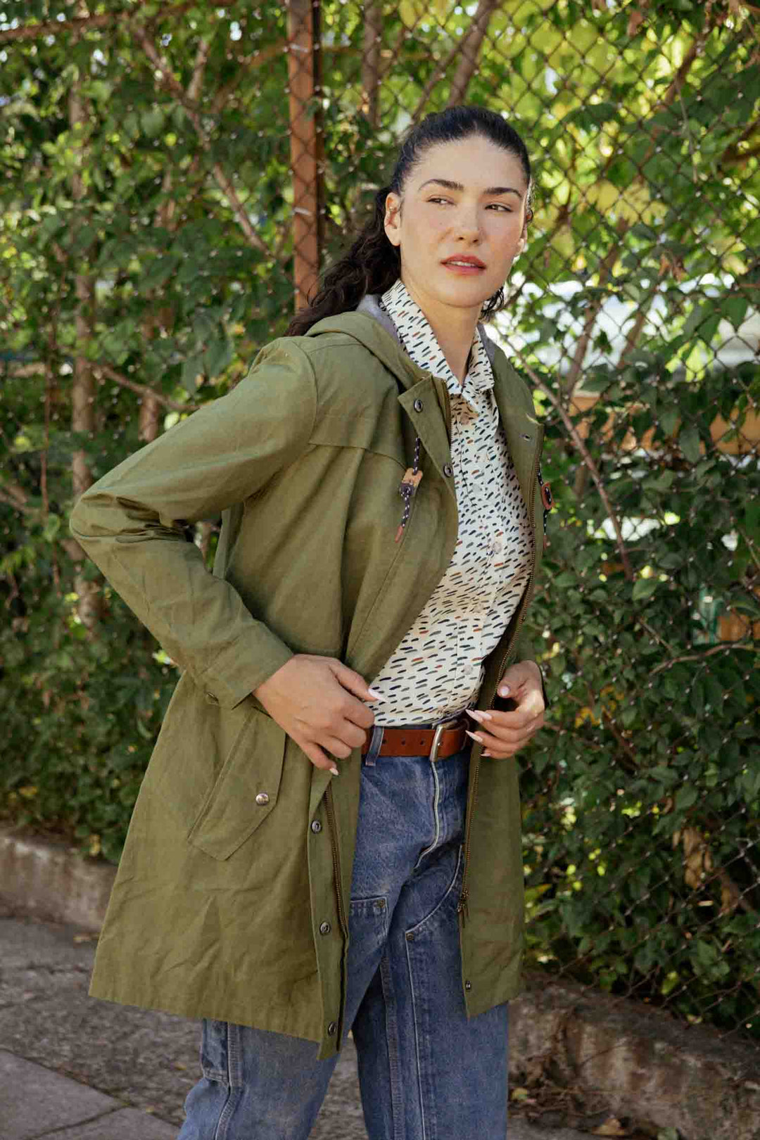 A woman stands outdoors in front of greenery, wearing the Bridge & Burn Briar Jacket in olive waxed canvas over a patterned shirt, blue jeans, and a brown belt. She looks to the side with one hand on her jacket.
