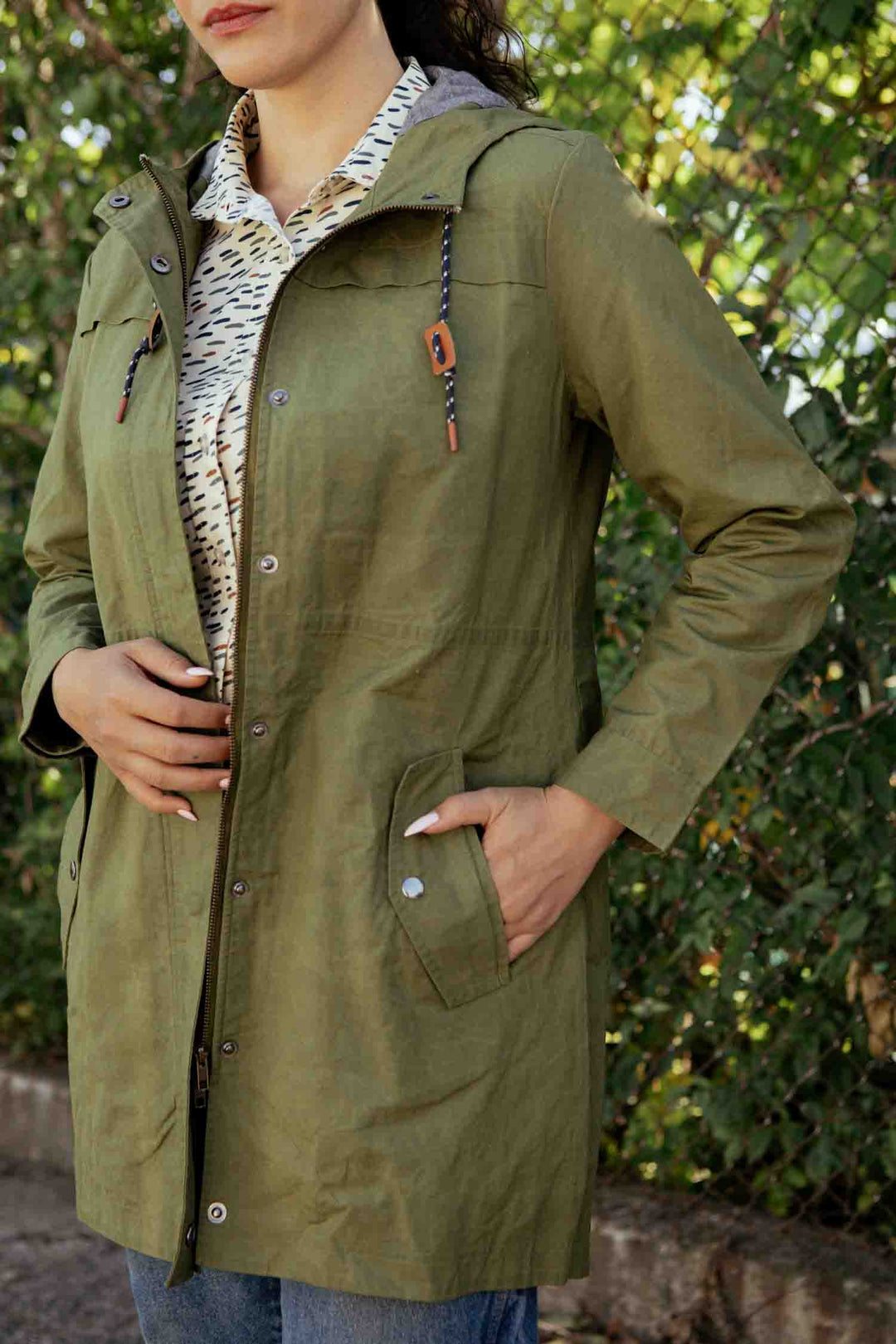 A woman stands outdoors in the Bridge & Burn Briar Jacket in Olive Waxed Canvas over a patterned shirt, her right hand on her stomach and left in a pocket, framed by greenery and a fence.