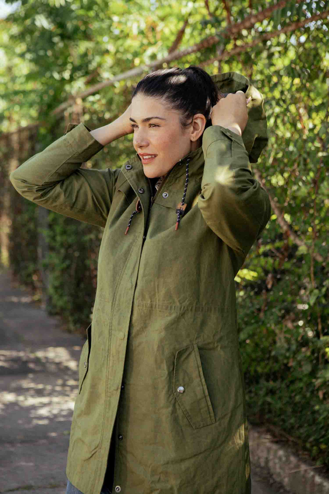 A person outdoors adjusts the hood of their Bridge & Burn Briar Jacket in Olive Waxed Canvas. Sunlight and green foliage in the background highlight their rain-ready style.