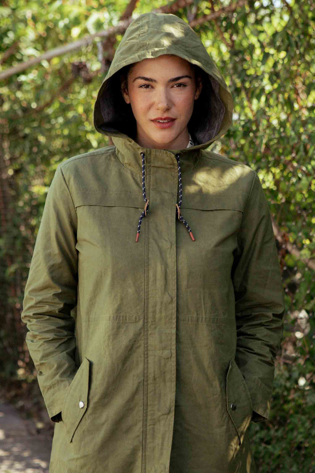 A person stands outdoors in the rain-ready Briar Jacket in Olive Waxed Canvas by Bridge & Burn, hands in pockets, with lush green foliage in the background.