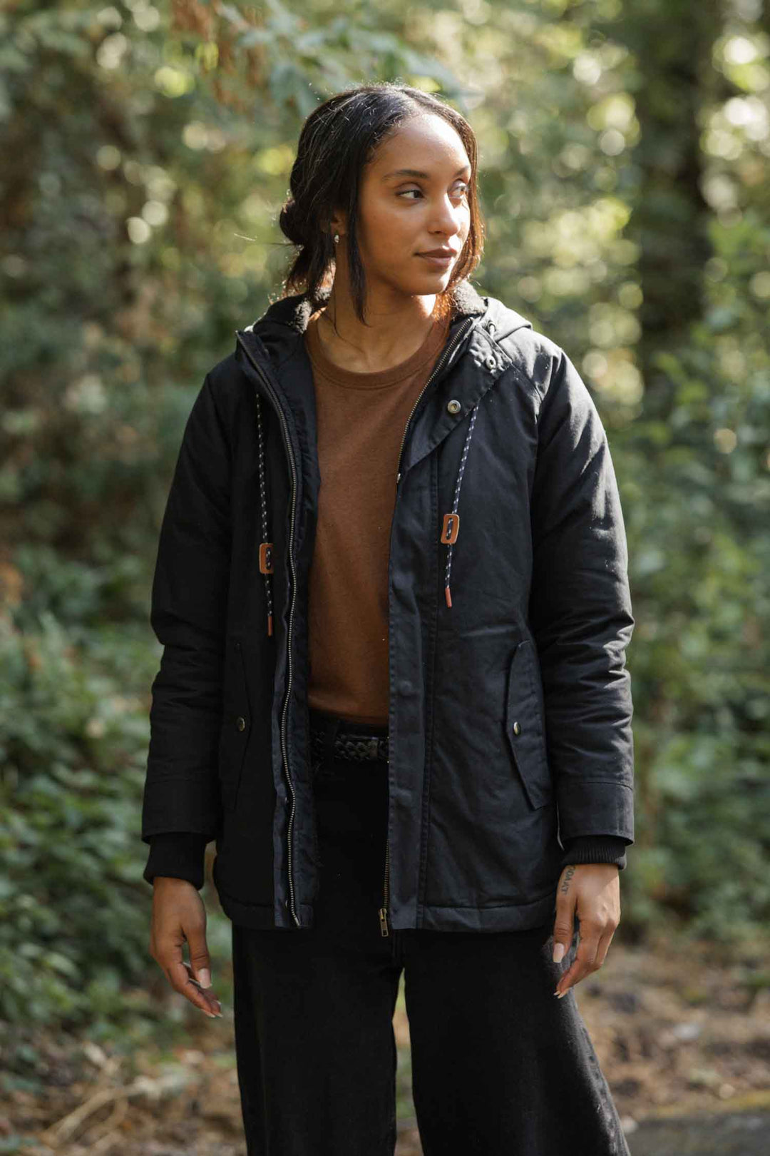 A woman with wavy hair stands outdoors in sunlight, wearing the Bridge & Burn Sequoia Parka in Black Waxed Canvas over a brown shirt, her hands relaxed at her sides as she looks slightly to the right among wooded trees.