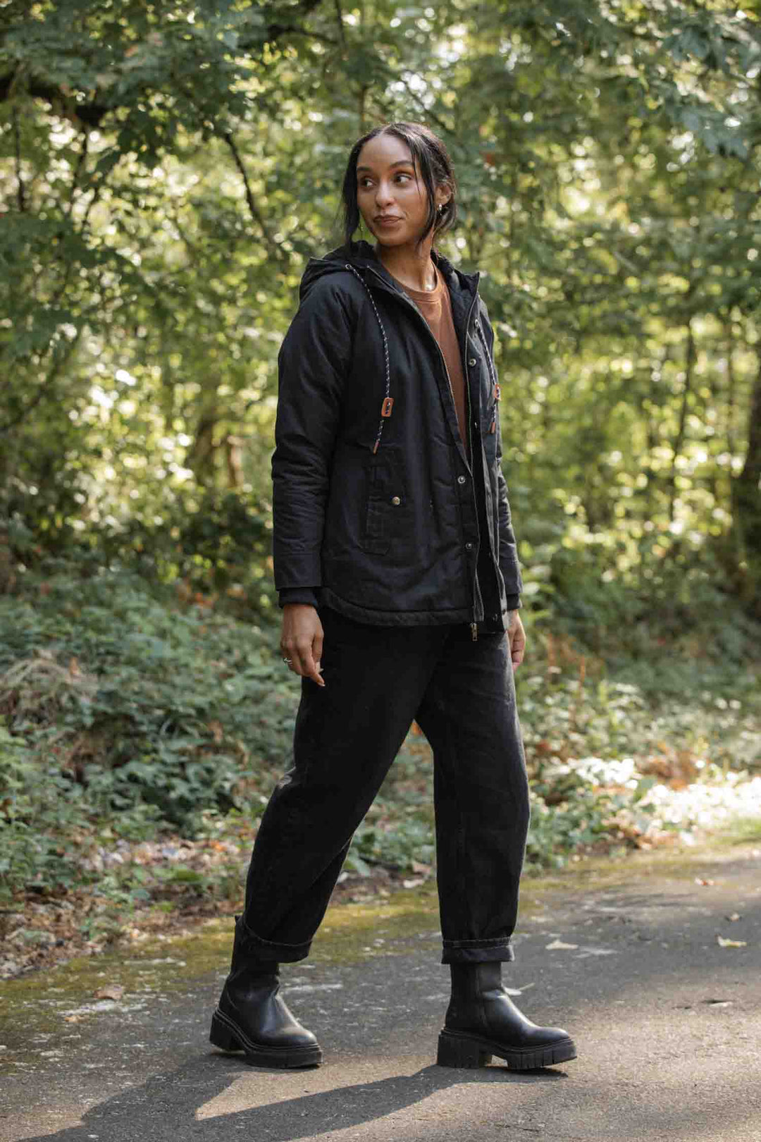 A woman stands on a shaded forest path, wearing the Bridge & Burn Sequoia Parka in black waxed canvas, paired with black pants and boots. She gazes calmly to the side, surrounded by lush green trees and foliage.