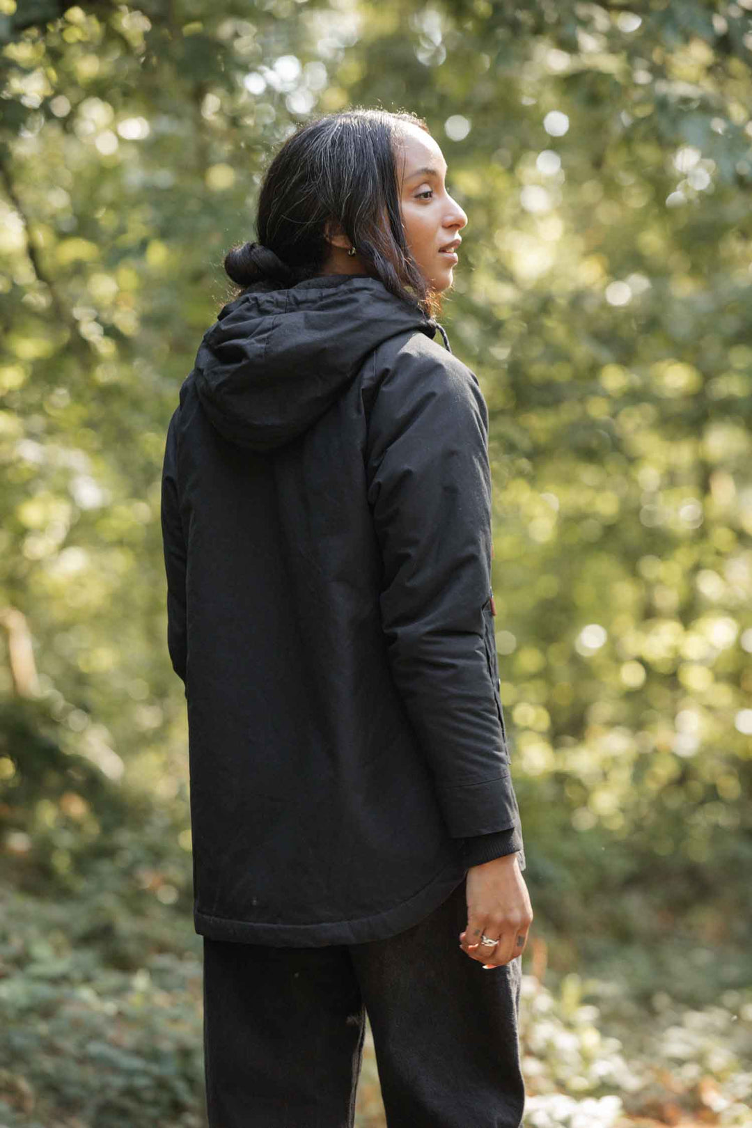 A person with long dark hair, wearing the Bridge & Burn Sequoia Parka in Black Waxed Canvas, stands outdoors in a sunlit green forest, looking over their shoulder.