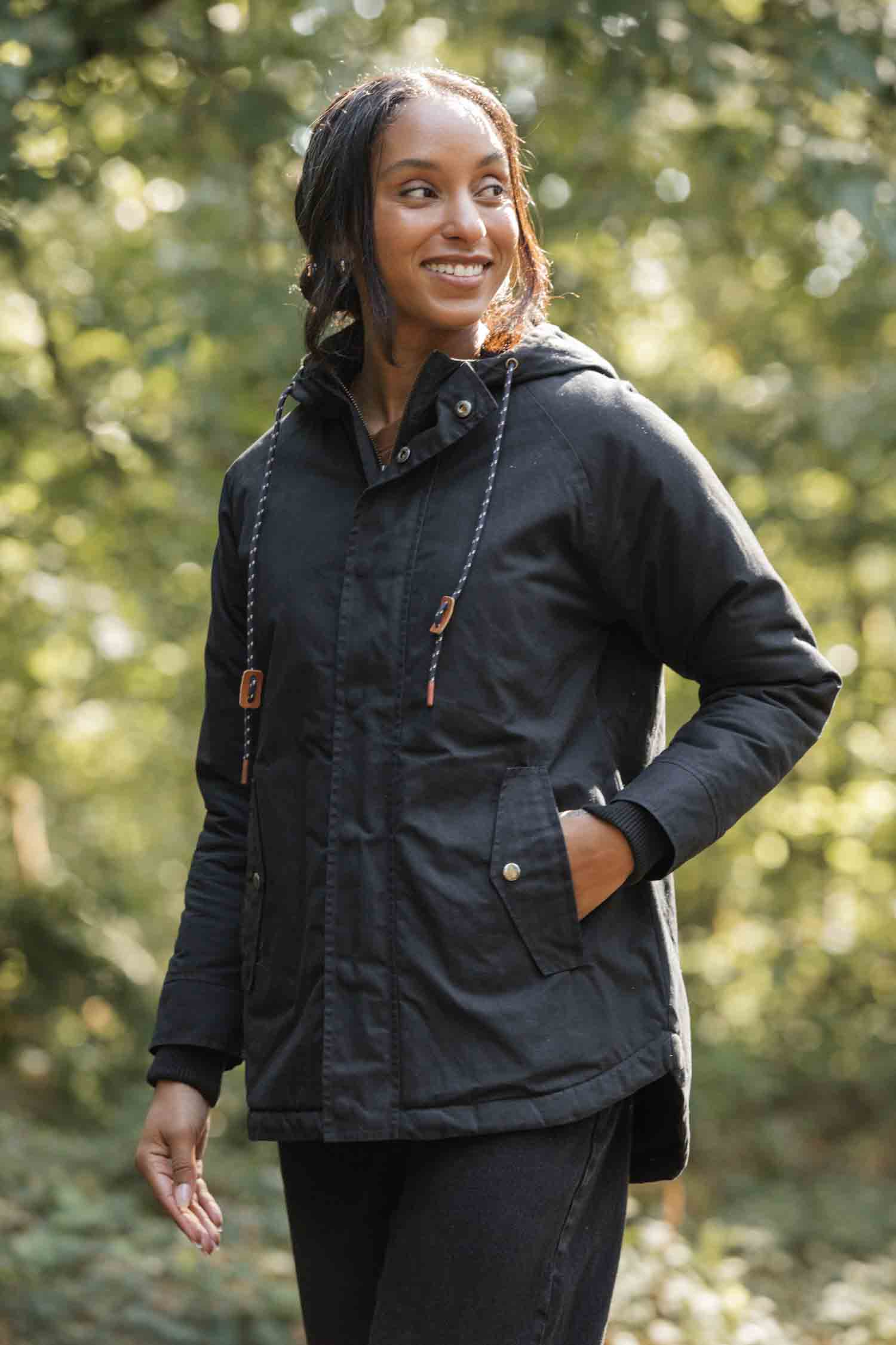 A woman wearing the Bridge & Burn Sequoia Parka in black waxed canvas stands outdoors in a sunlit green forest, smiling and looking to the side with one hand in her pocket.