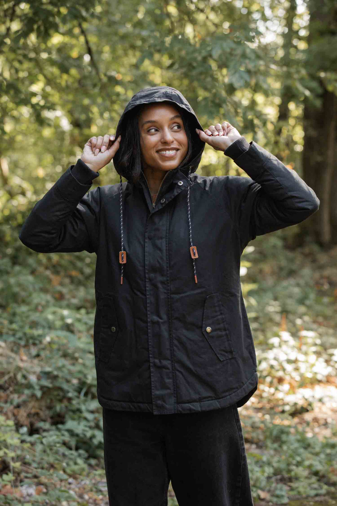 A woman stands in a wooded area, smiling and holding the hood of her Bridge & Burn Sequoia Parka in Black Waxed Canvas. She wears dark pants and is surrounded by green foliage.