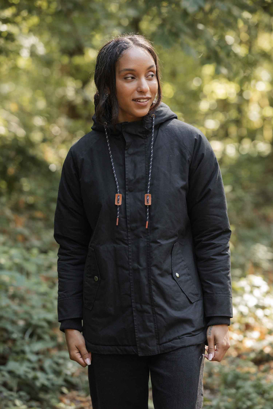 A woman stands outdoors among green foliage, smiling gently. She wears the Bridge & Burn Sequoia Parka in black waxed canvas, showcasing its stylish design and cozy fleece lining.