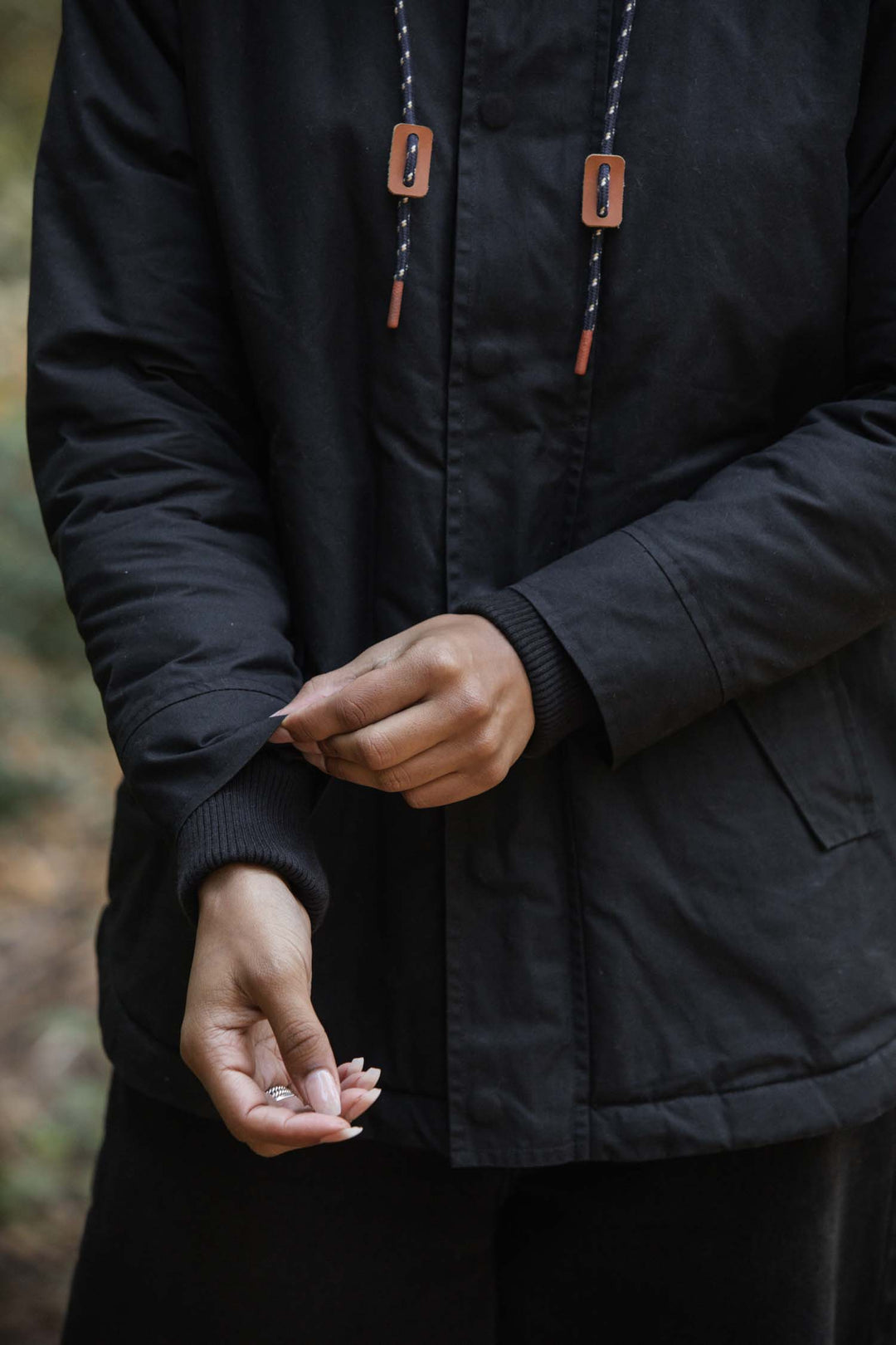 A person wearing the Bridge & Burn Sequoia Parka in black waxed canvas adjusts their sleeve with one hand while holding a small item in the other. The background is blurred and outdoors.