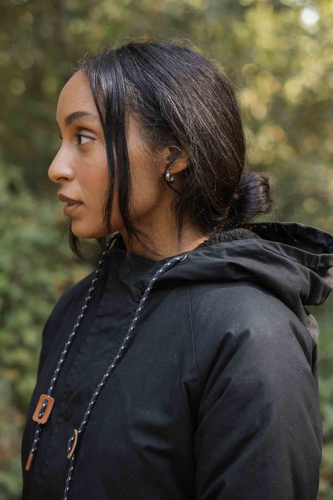 A woman with long dark hair tied back stands outdoors in profile against a green, blurred background, wearing the Bridge & Burn Sequoia Parka in black waxed canvas.