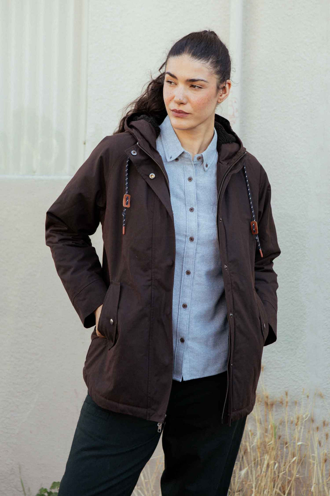 A person with long dark hair pulled back is wearing the Bridge & Burn Sequoia Parka in Umber Waxed Canvas over a light gray button-up shirt, standing outdoors near a light wall and dry grass.