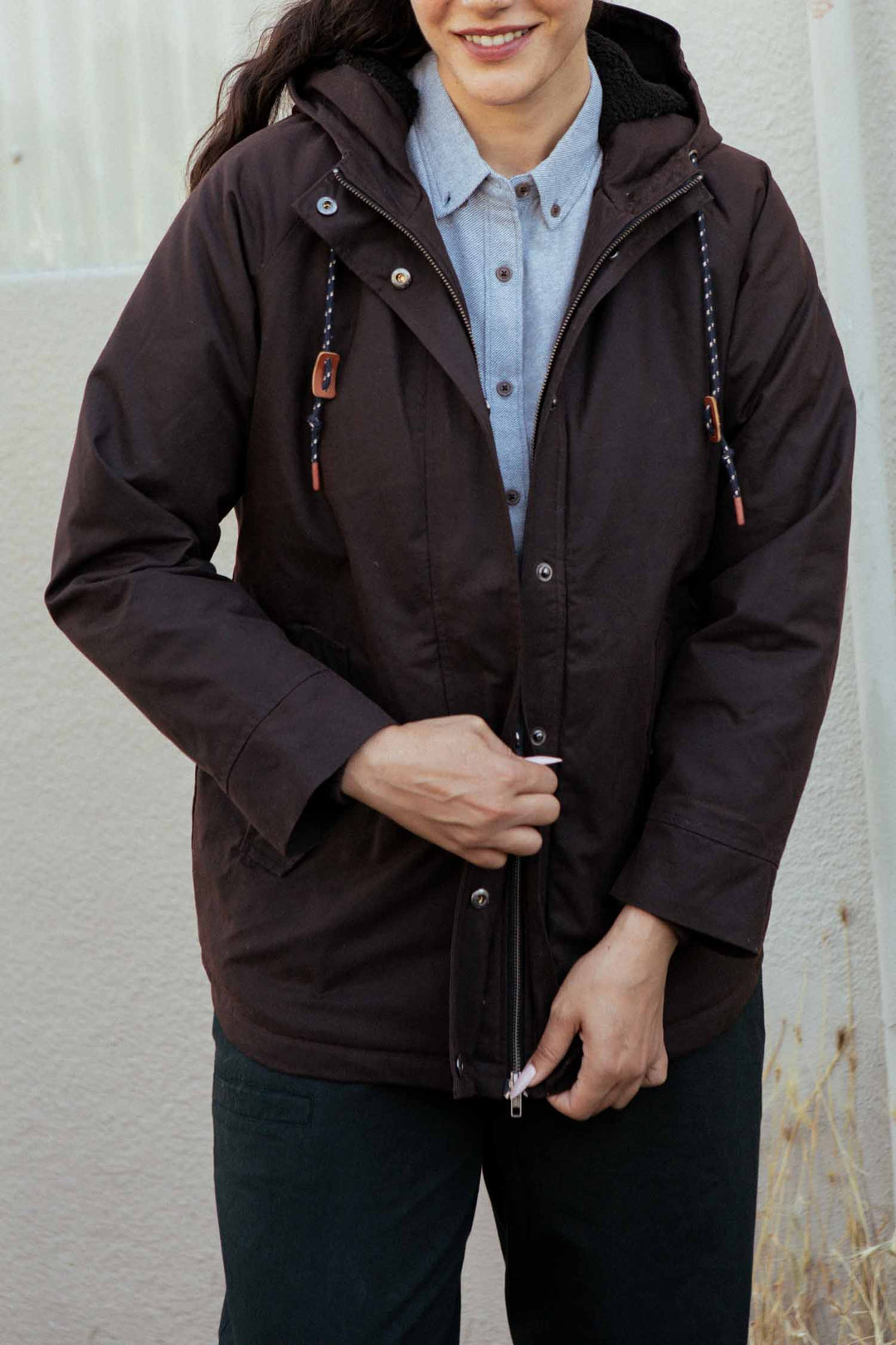 A woman smiles and zips up the Bridge & Burn Sequoia Parka in umber waxed canvas over a light gray button-up shirt, standing outdoors against a light-colored wall.