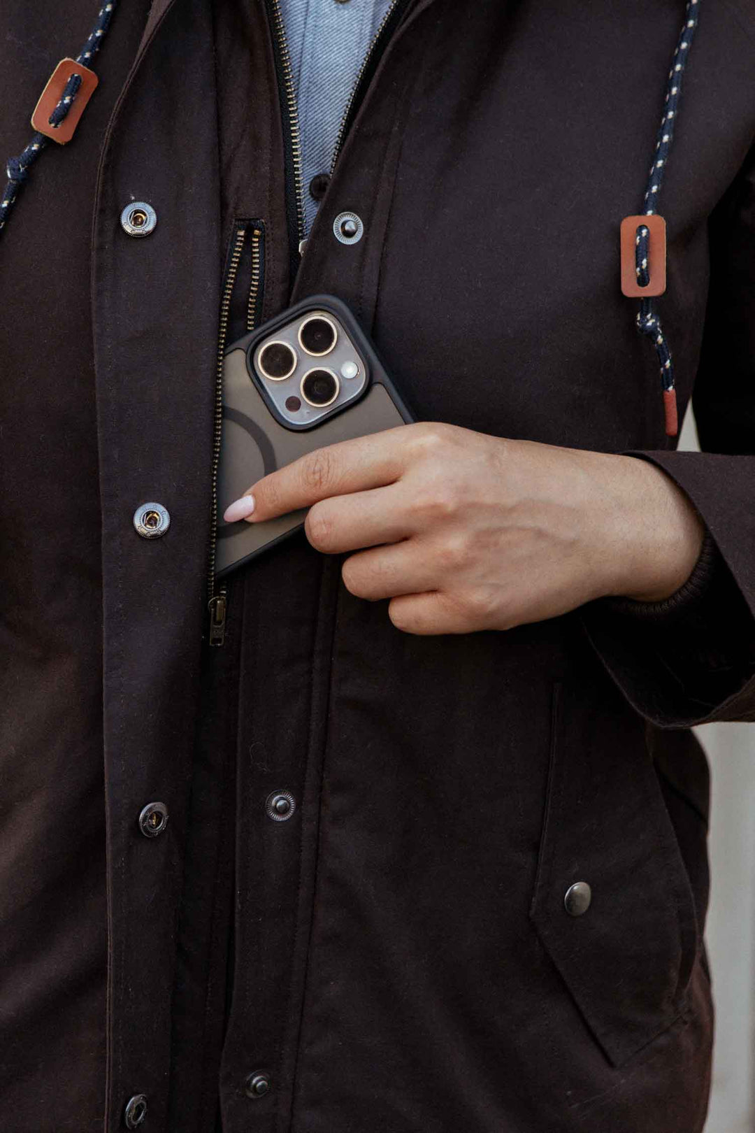 A person wearing the Bridge & Burn Sequoia Parka in umber waxed canvas unzips their chest pocket to place a patterned-cased smartphone inside; only their hand and torso are visible.