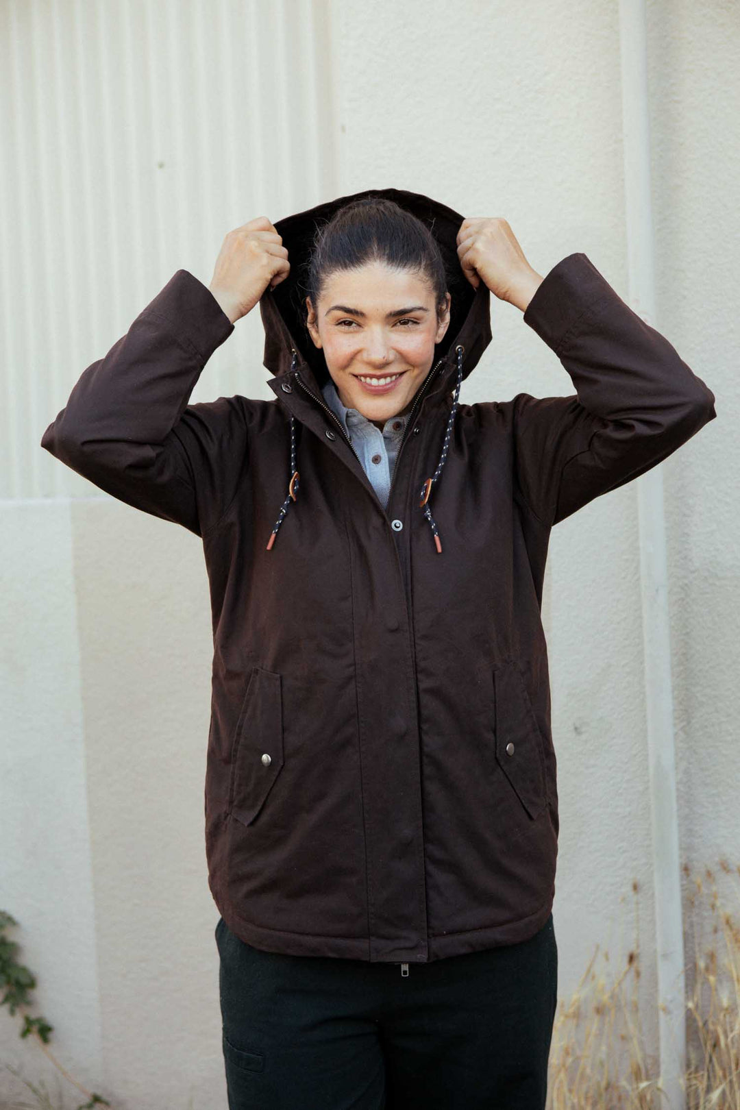 A person stands smiling outdoors, holding up the hood of the Bridge & Burn Sequoia Parka in Umber Waxed Canvas. They wear a light shirt underneath, with light walls and dry grass in the background.
