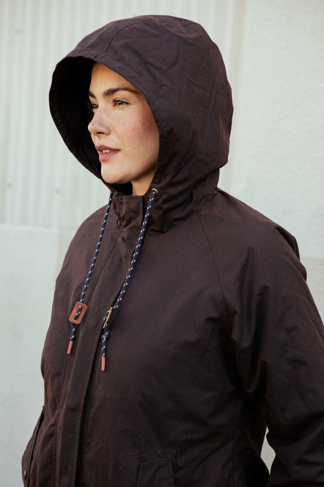A person wears the Bridge & Burn Sequoia Parka in umber waxed canvas, looking to the side against a light-colored wall with vertical lines. The hooded jacket features drawstrings for an adjustable fit.