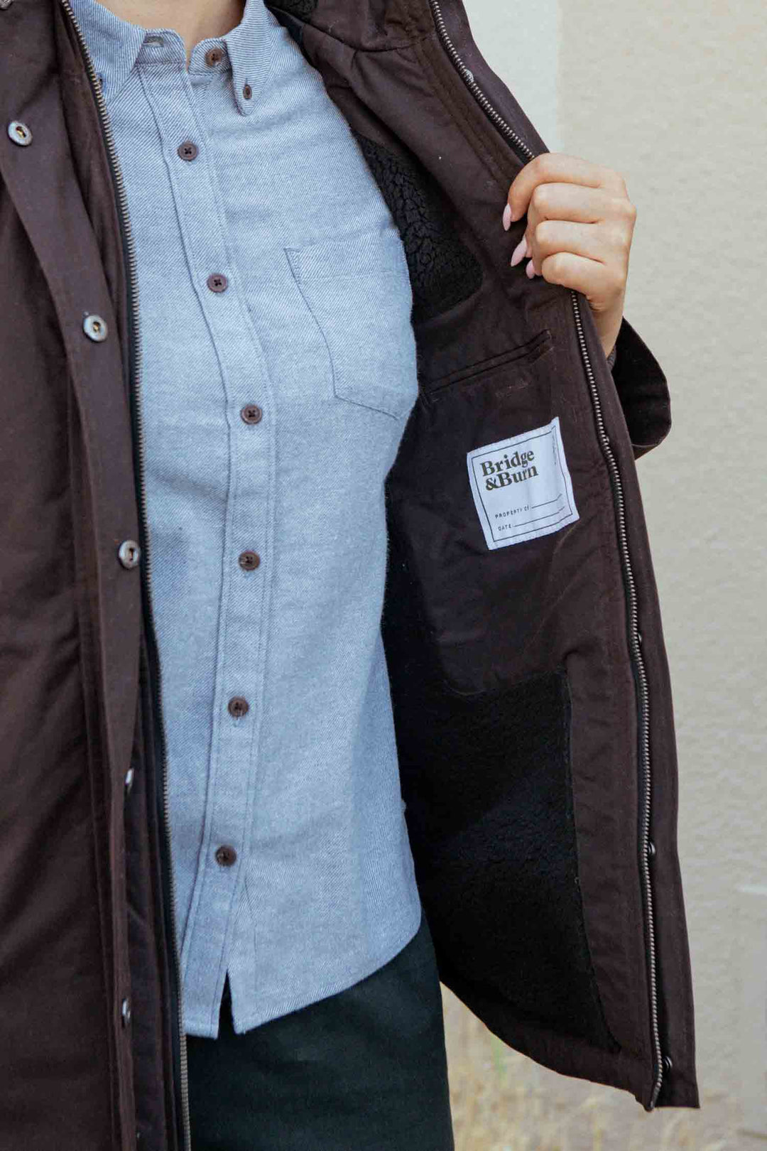 A person wearing a light blue shirt and the Bridge & Burn Sequoia Parka in Umber Waxed Canvas holds open the jacket to show the Bridge & Burn label sewn inside.