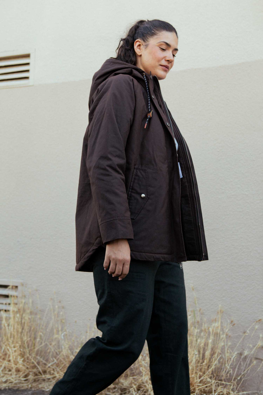 A person with dark hair in a ponytail wears the Bridge & Burn Sequoia Parka in Umber Waxed Canvas and dark pants, walking outdoors beside a beige wall and dry grass.