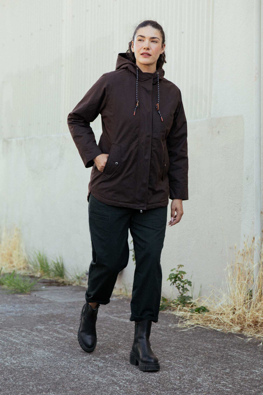 A person walks outdoors along a paved path, wearing the Bridge & Burn Sequoia Parka in umber waxed canvas, black pants, and black boots. The background features a light-colored wall and dry grass.