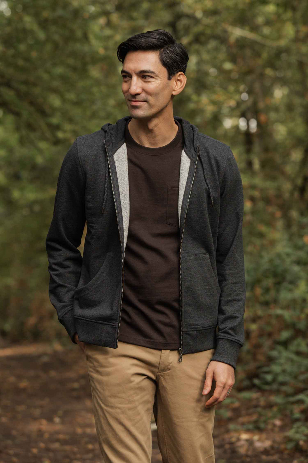 A man stands on a wooded path, smiling and looking to the side, wearing Bridge & Burn's Ridge Hoodie in Graphite, paired with a brown T-shirt and khaki pants. Lush green trees fill the background.