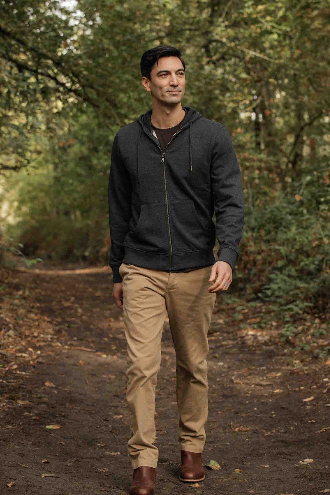 A man wearing the Bridge & Burn Ridge Hoodie in Graphite, beige pants, and brown boots walks along a dirt path in a forest surrounded by green trees.