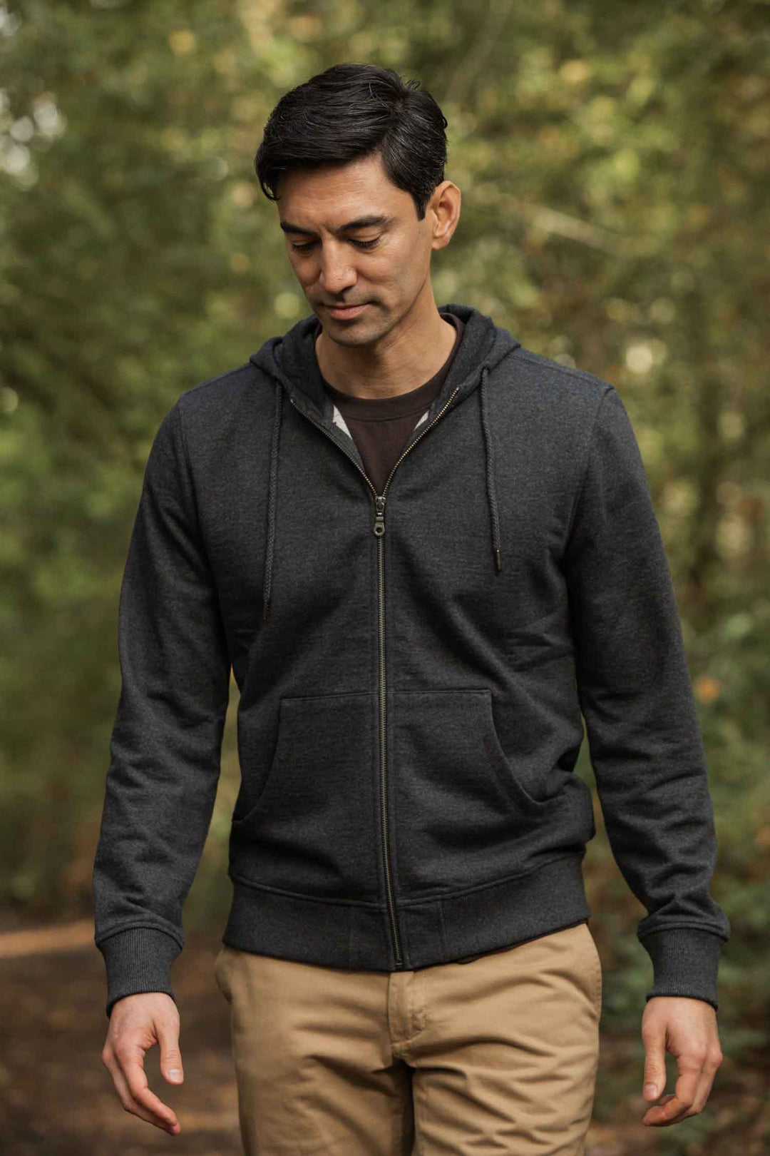 A man in the Bridge & Burn Ridge Hoodie in Graphite and tan pants walks on a wooded path, looking down with a neutral expression. The blurred green background features trees.
