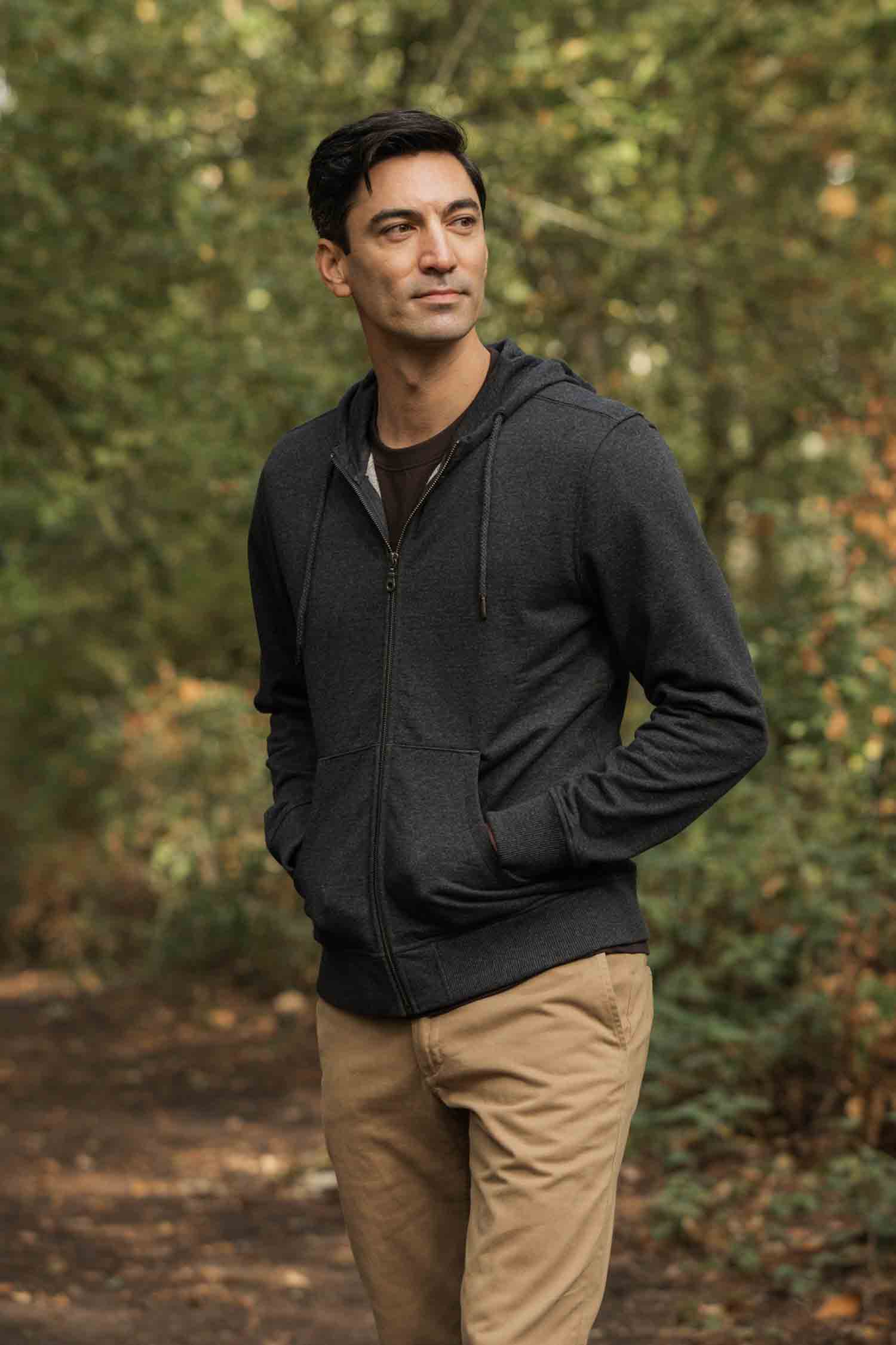 A man with short dark hair stands on a wooded path, wearing the Bridge & Burn Ridge Hoodie in Graphite—made from recycled fibers—and tan pants, hands in his pockets, surrounded by green trees and foliage.