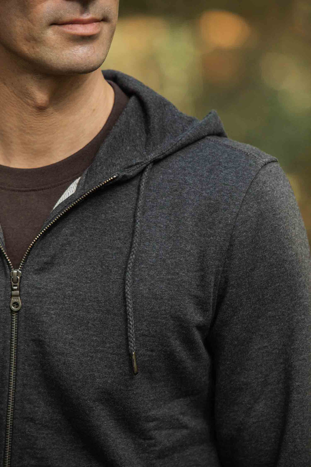 A man stands outdoors in a Bridge & Burn Ridge Hoodie in Graphite, layered over a brown T-shirt. Made from recycled fibers, the zip-up hoodie is dark gray. His partially visible face contrasts with the softly blurred warm background.