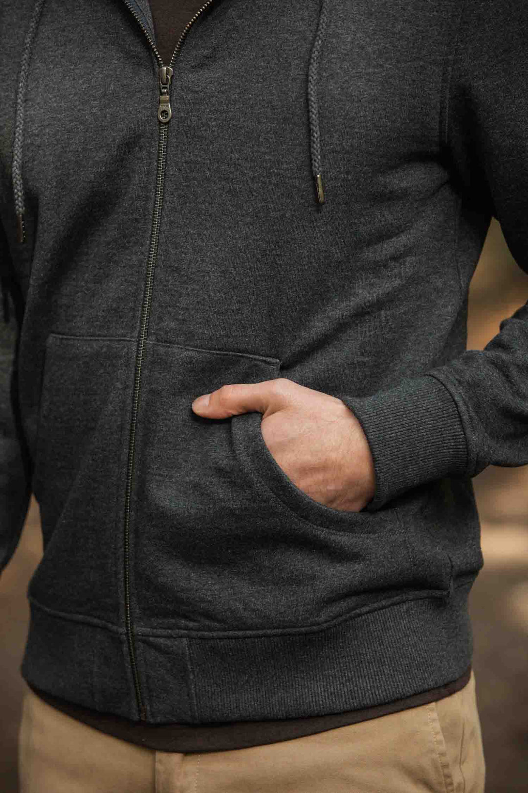 A person wears the Bridge & Burn Ridge Hoodie in Graphite with tan pants, standing outdoors with one hand in the hoodie’s front pocket. Only the torso and part of one arm are visible.