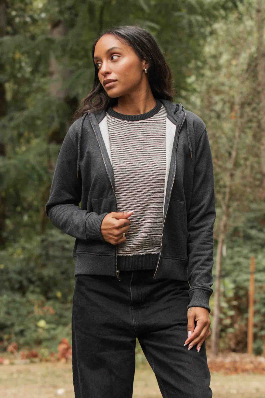 A woman stands outdoors in front of greenery, wearing the Bridge & Burn Mason Hoodie in Graphite over a striped sweater and black pants. She looks to the side, holding her jacket with one hand.