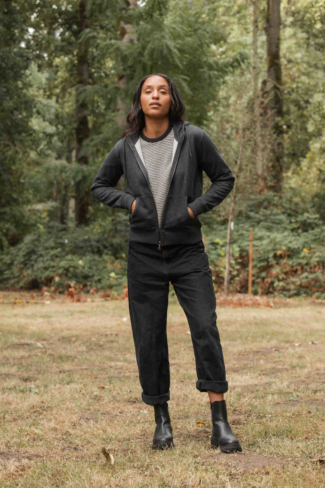 A person stands outdoors on grass, hands in the pockets of the Bridge & Burn Mason Hoodie in Graphite, paired with black cuffed pants, a striped shirt, and black boots. Trees and greenery fill the background.