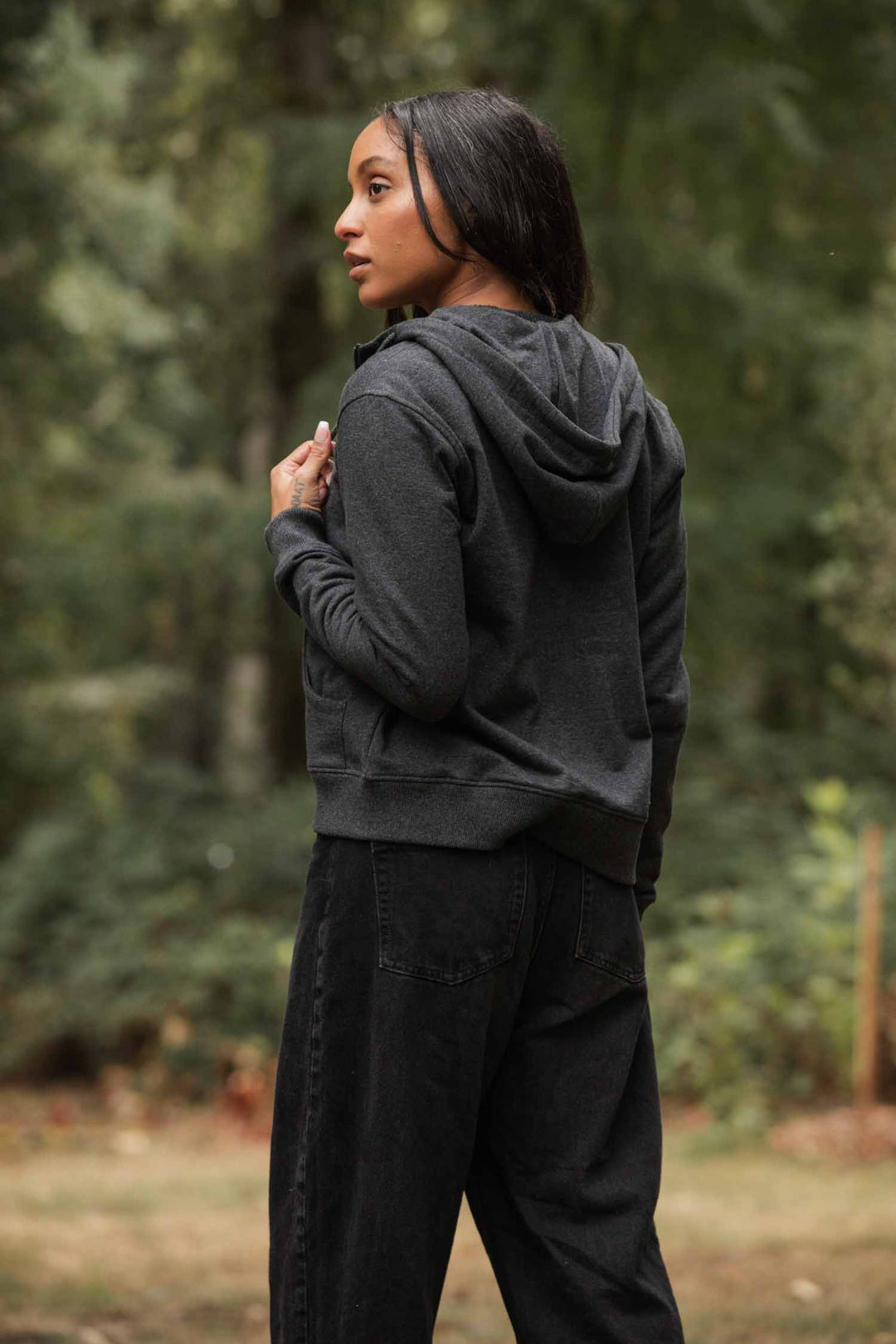 A woman with long dark hair stands outdoors in a forest, wearing the Bridge & Burn Mason Hoodie in Graphite and black pants, gazing off to the side.
