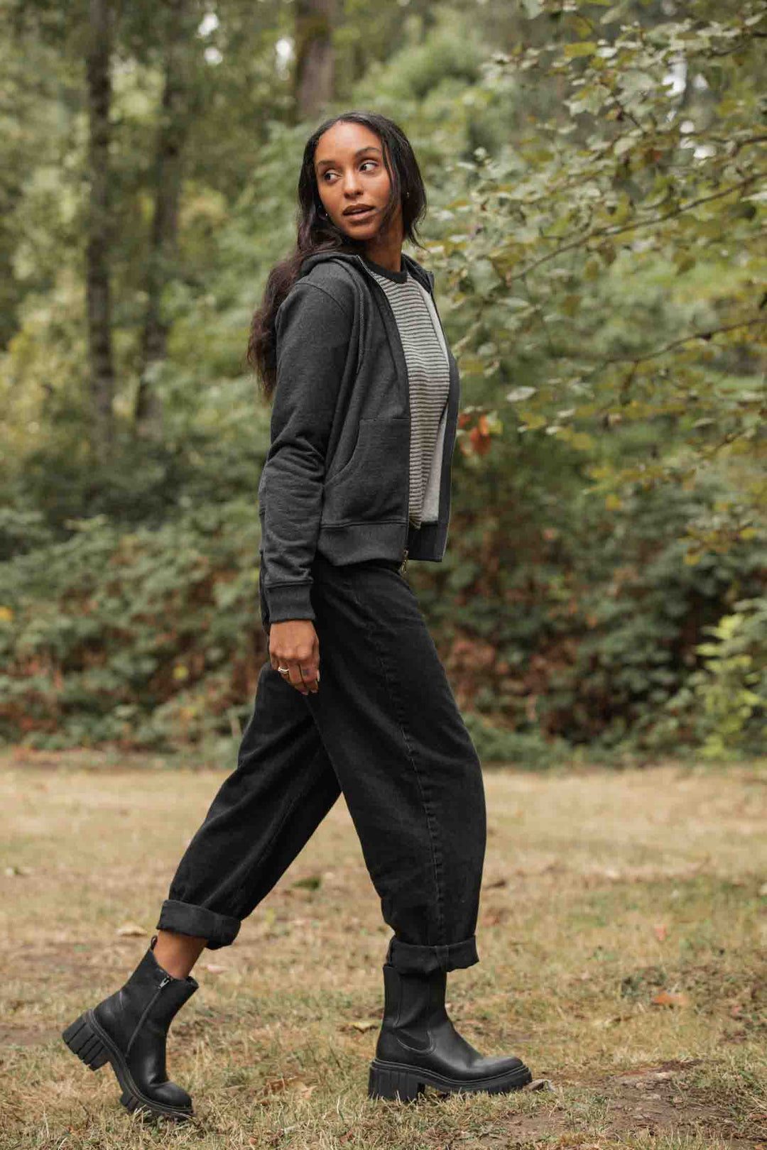 A woman with long hair walks outdoors on grass, wearing the Bridge & Burn Mason Hoodie in Graphite, a striped shirt, black pants, and black chunky boots, surrounded by green trees and foliage.