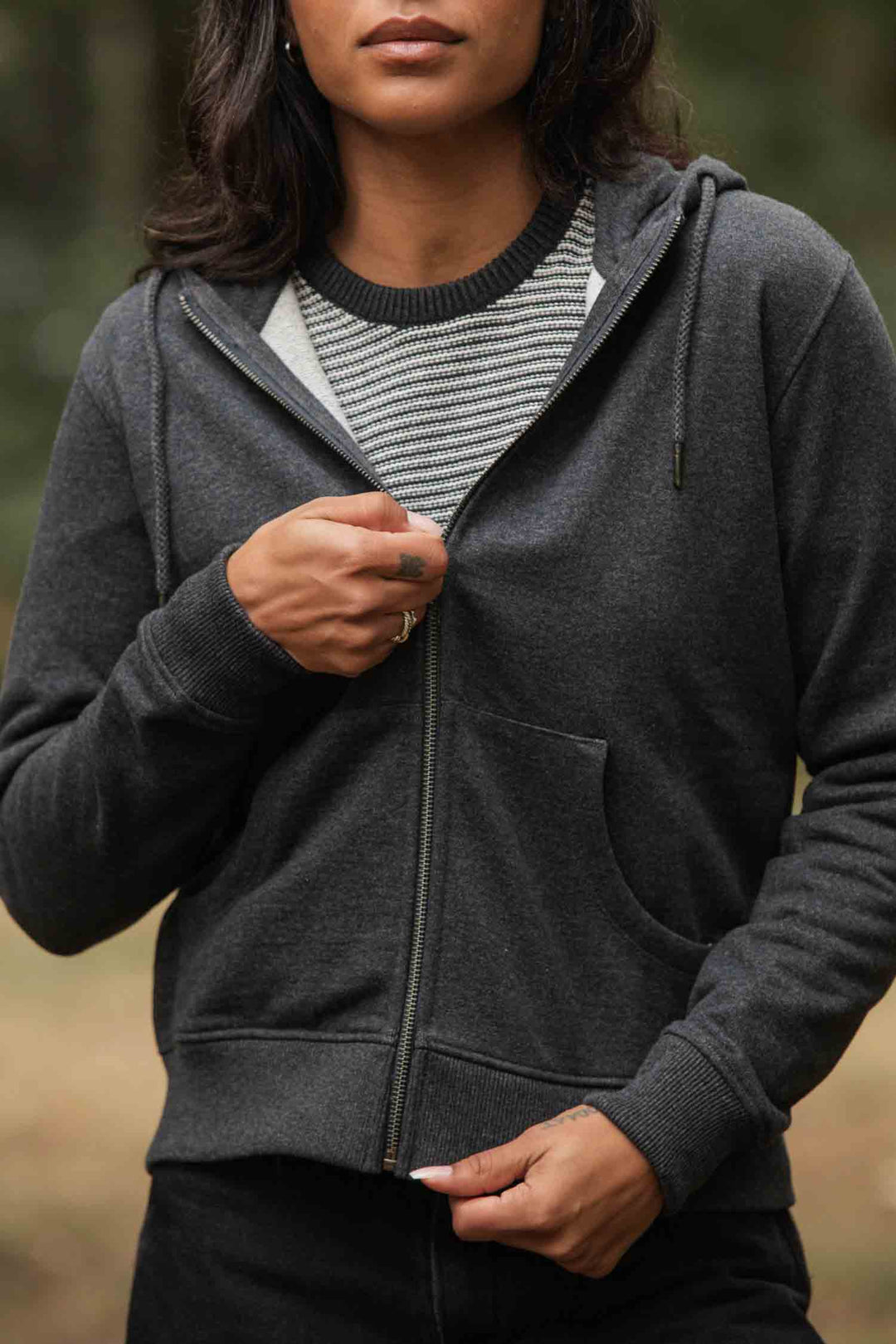 A person is partially unzipping the Bridge & Burn Mason Hoodie in Graphite, worn over a striped shirt. The blurred green background highlights their torso and hands, while their face is only partially visible.