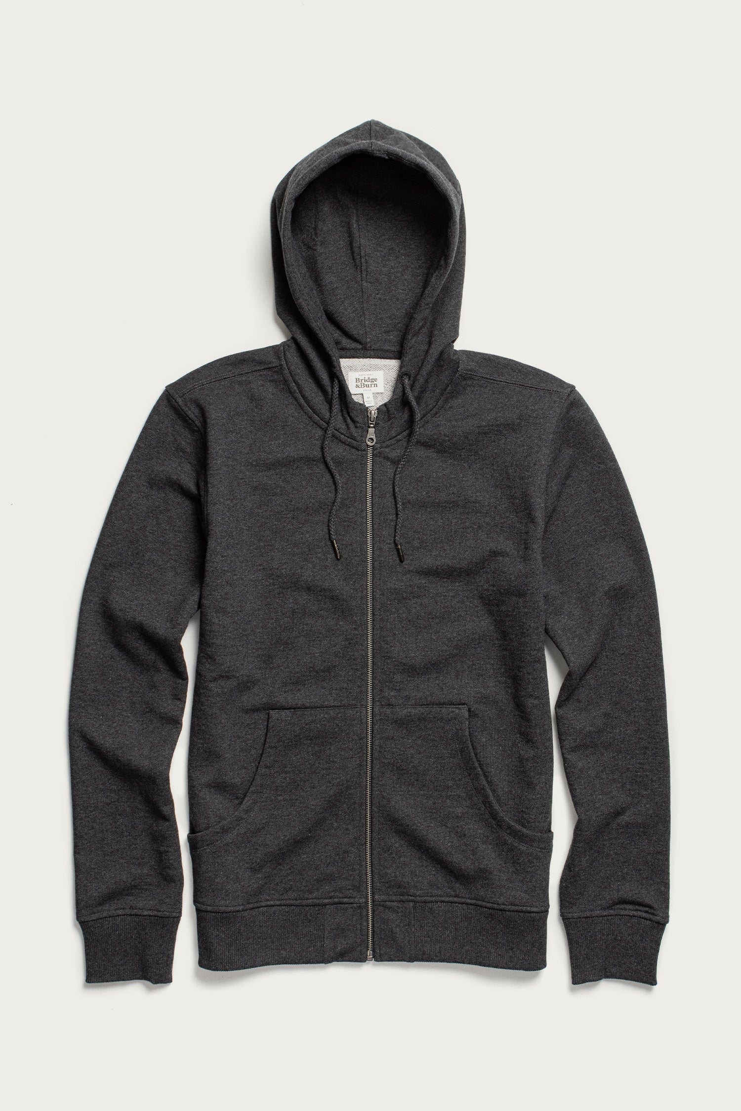 The Ridge Hoodie in Graphite by Bridge & Burn is a dark gray zip-up hoodie made from organic cotton, with a hood, drawstrings, front zipper, and two front pockets, shown laid flat on a white background.