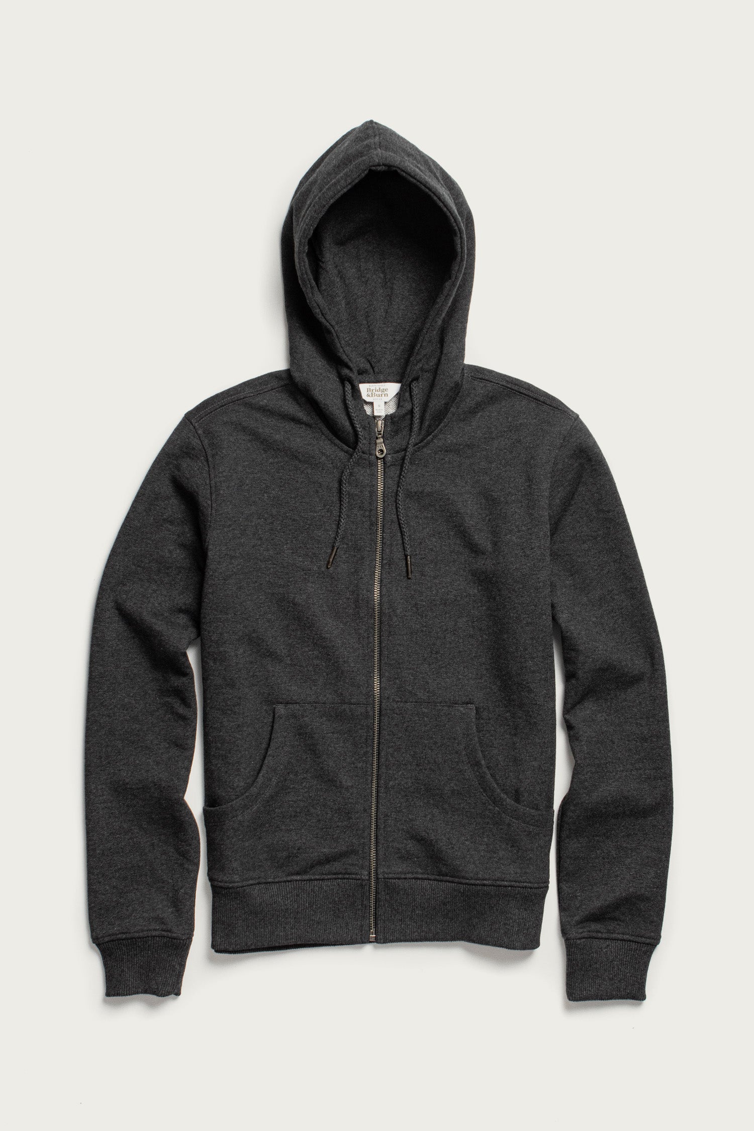 The Bridge & Burn Mason Hoodie in Graphite is a sustainable, dark gray French terry hoodie featuring a front zipper, two pockets, and an adjustable drawstring hood, shown laid flat against a light background.