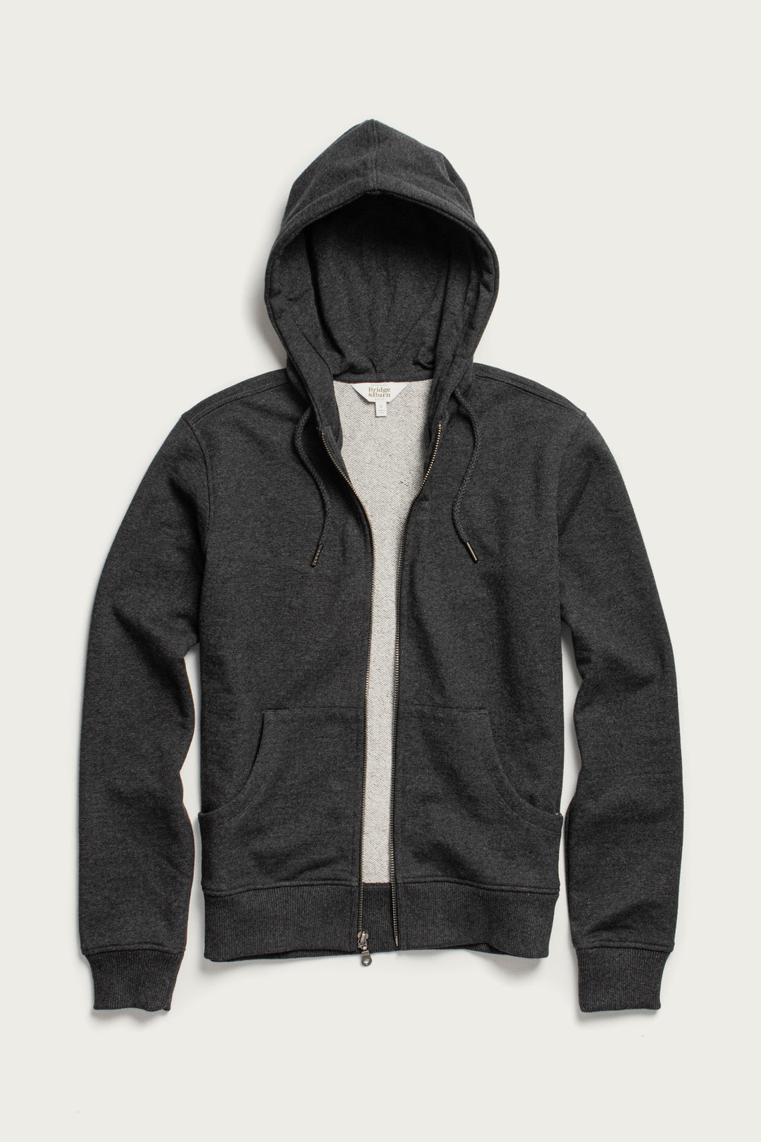 The Mason Hoodie in Graphite by Bridge & Burn is a dark gray French terry zip-up with front pockets, hood, drawstrings, and center zipper, shown flat on a light background.