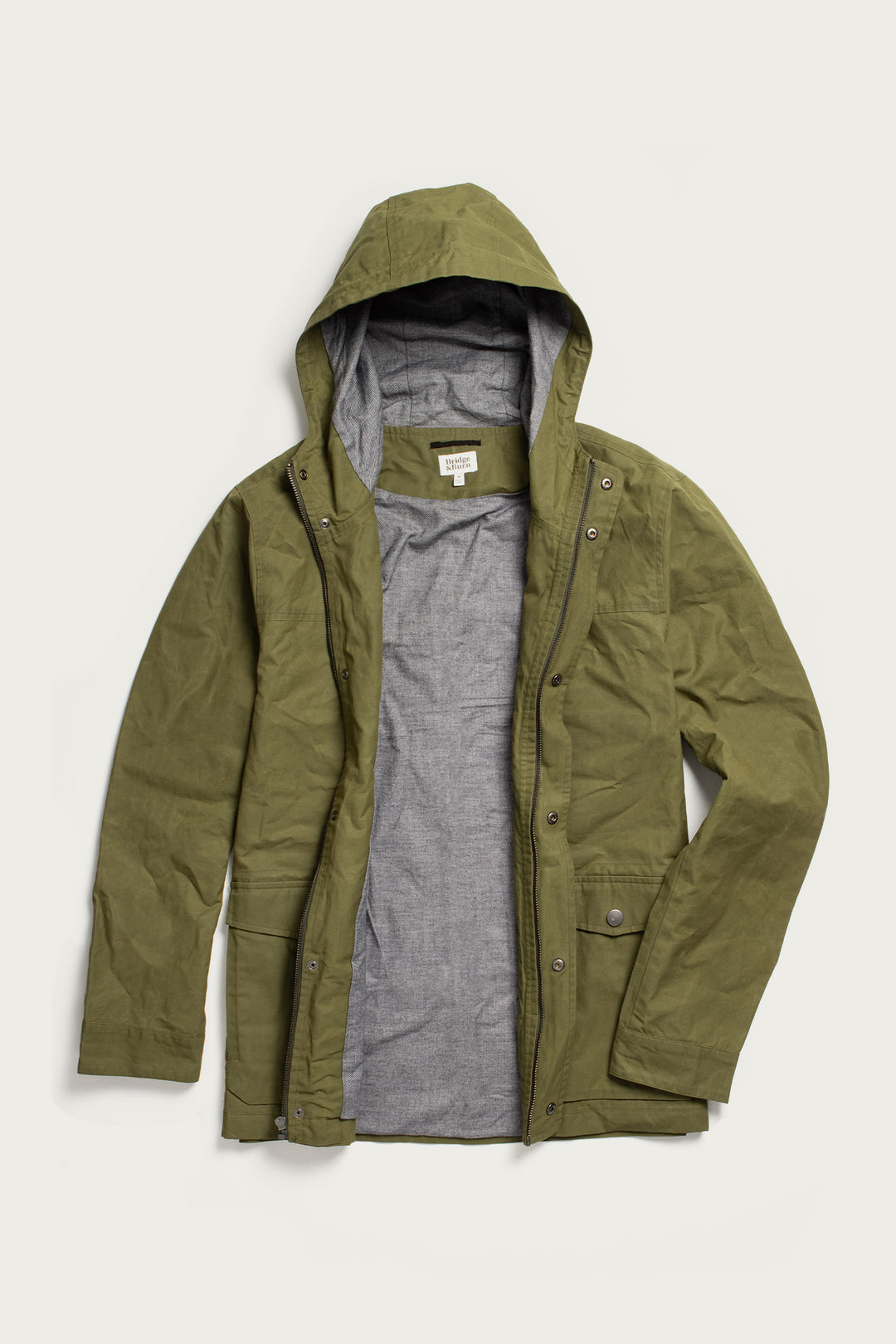 The Bridge & Burn Marshall Jacket in olive waxed canvas is a water-resistant, hooded jacket with a gray lining, snap buttons, and front pockets, shown unzipped and laid flat on a white background.