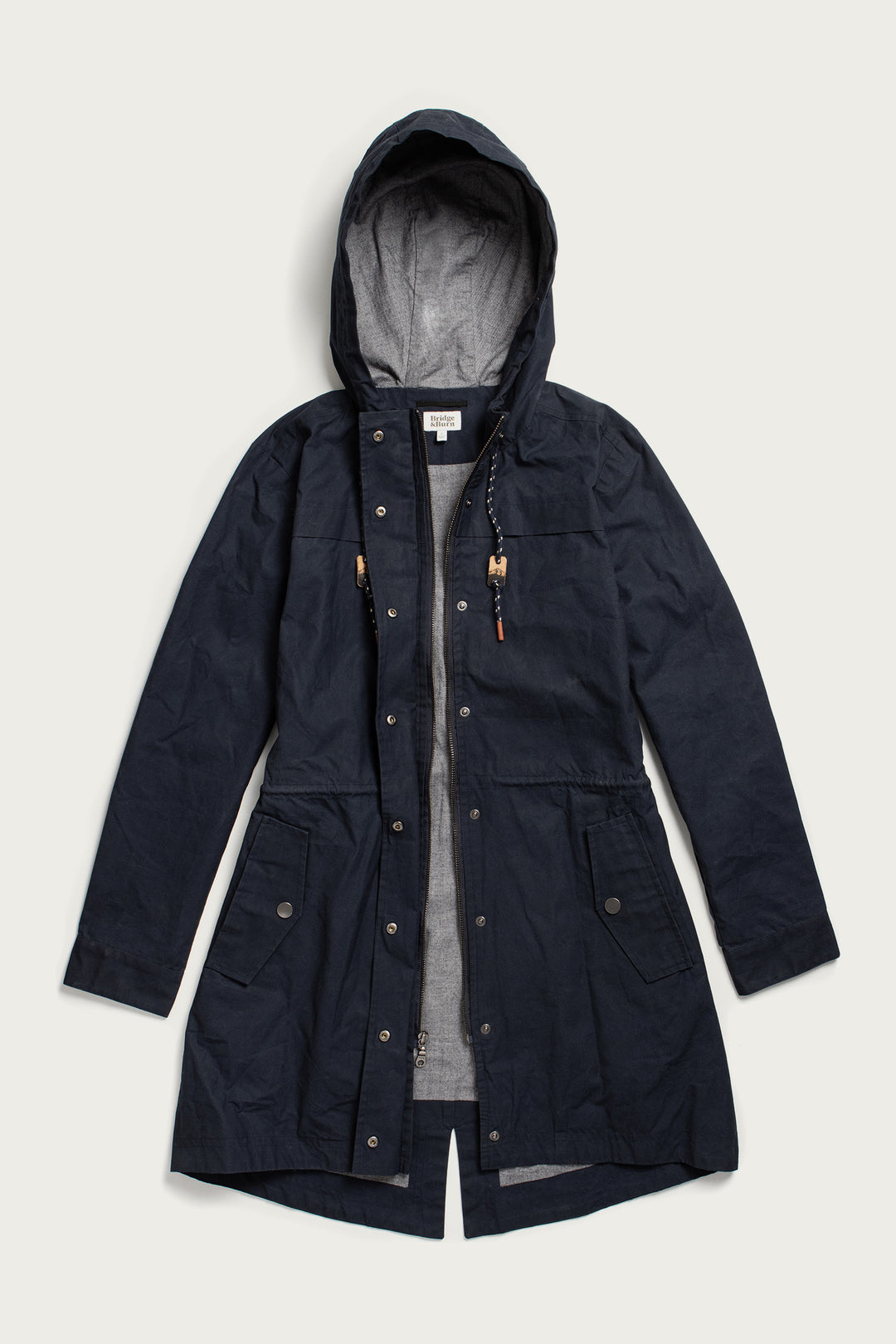 The Briar Jacket by Bridge & Burn is a navy waxed canvas parka with a hood, front zipper and snaps, two front flap pockets, adjustable waist and neck drawstrings, and a light gray lining—shown laid flat on a white background.