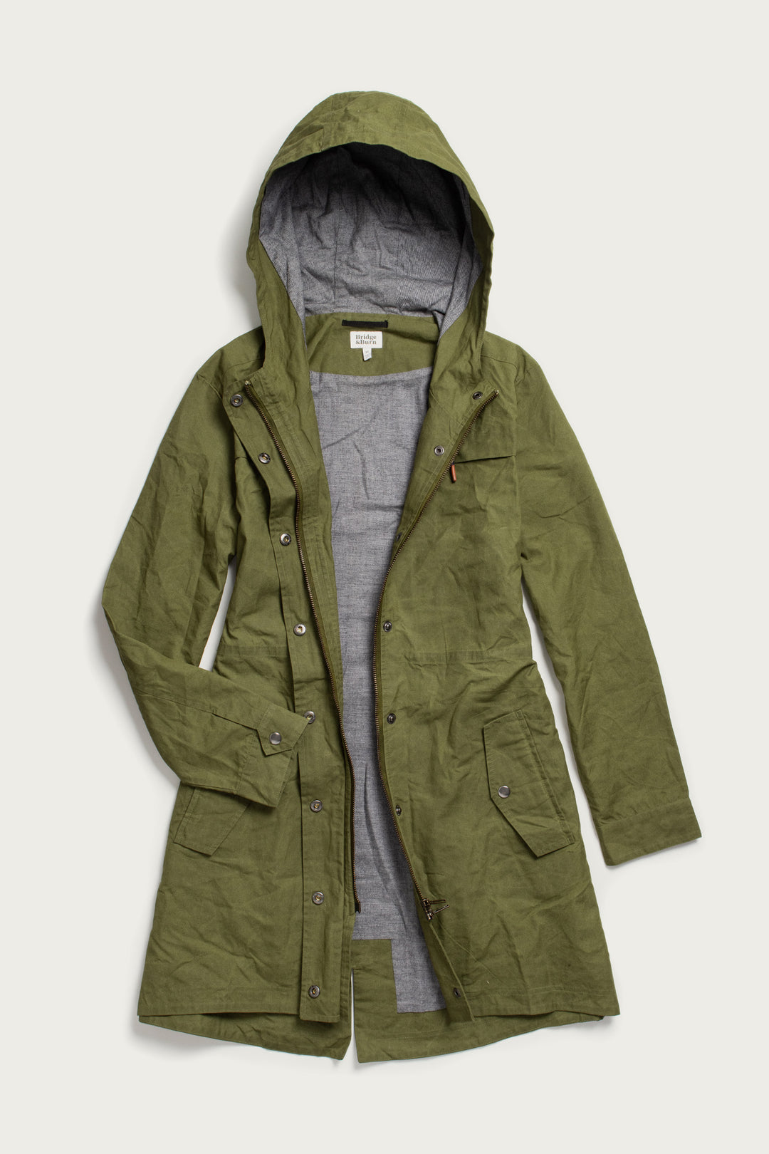 The Bridge & Burn Briar Jacket in Olive Waxed Canvas features British Millerain fabric, a hood, front snap buttons, two spacious front pockets, and a gray lining. Shown laid flat on a light background.