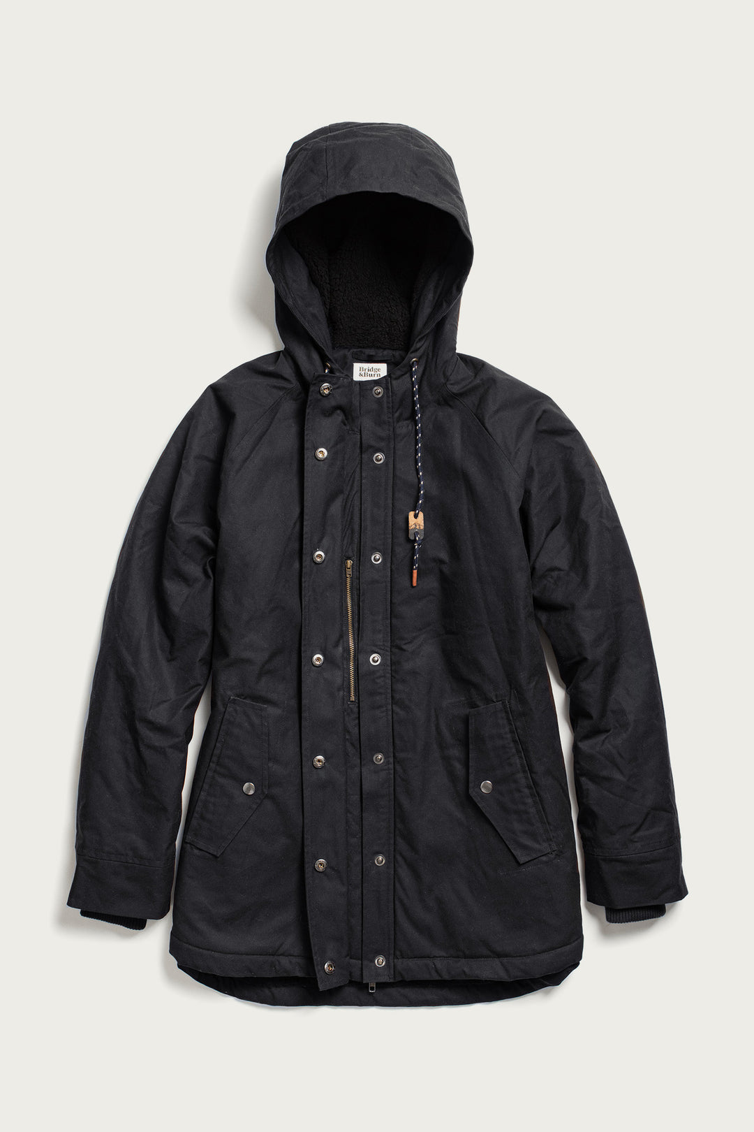 The Bridge & Burn Sequoia Parka in black waxed canvas features a front zipper with snap button closure, fleece lining for warmth, two front flap pockets, and a drawstring hood. Displayed on a plain light background.