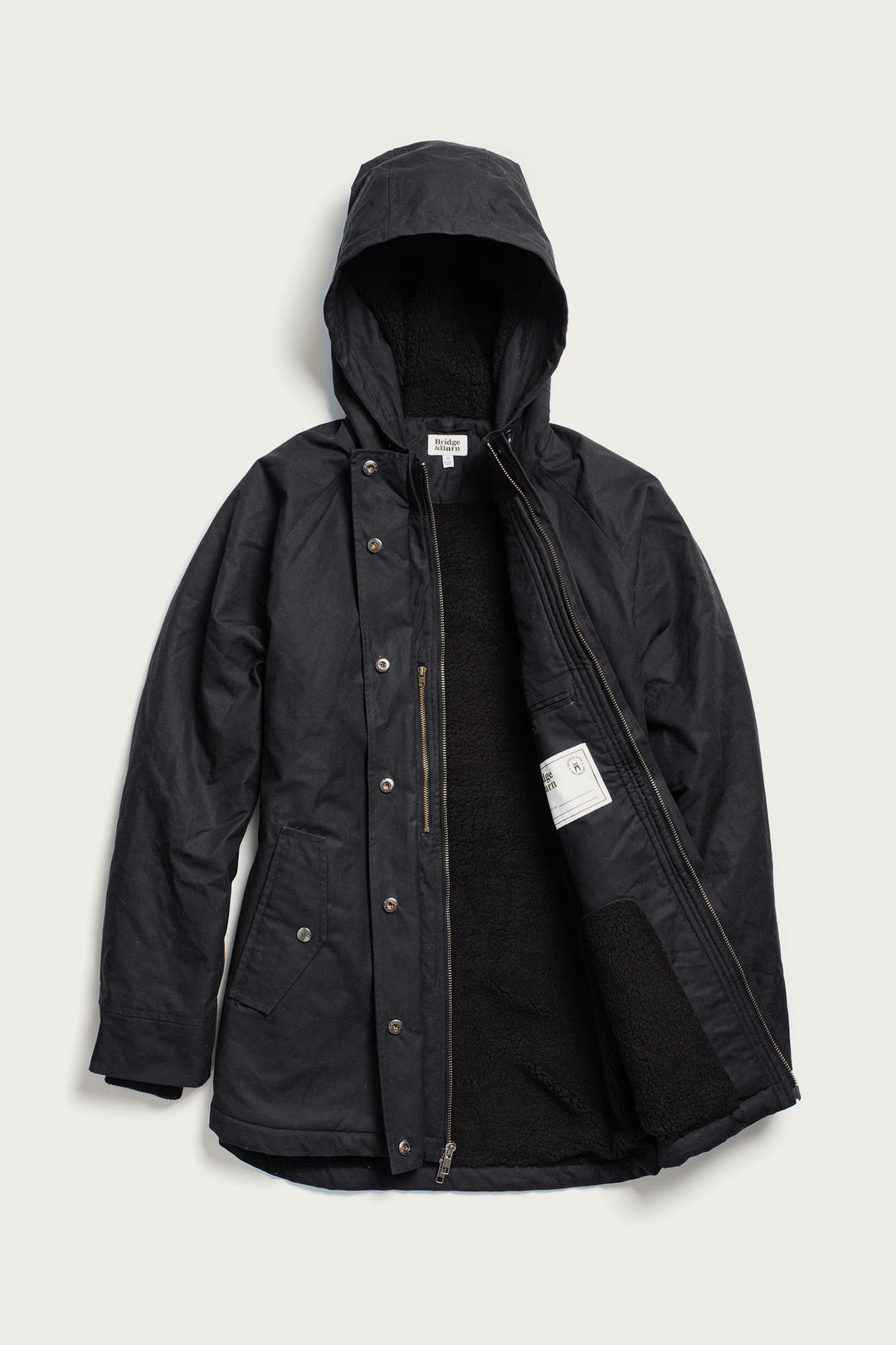 The Sequoia Parka by Bridge & Burn in black waxed canvas features a hood, snap buttons, zippered chest pocket, soft black fleece lining partially visible at the front, and a white label. The jacket is laid flat on a light background.