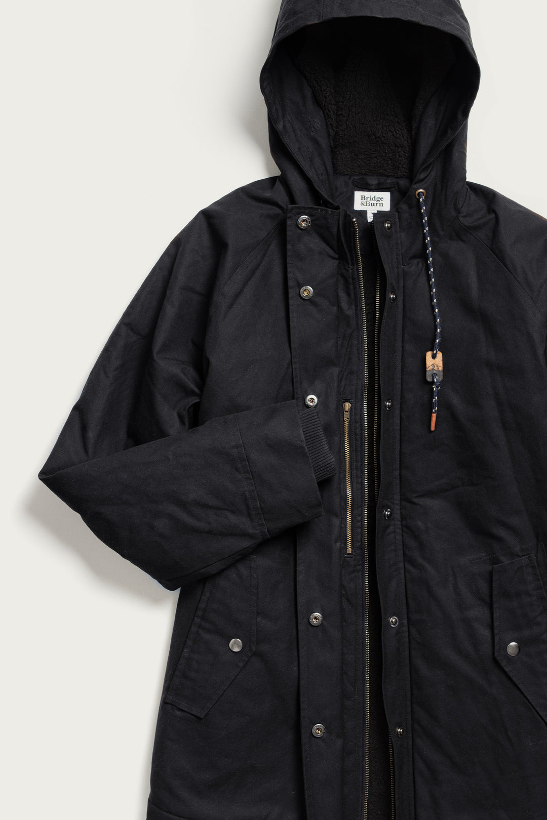 The Bridge & Burn Sequoia Parka in black waxed canvas features snap buttons, a zipper closure, side pockets, and a hood with a braided drawstring and wooden bead, shown on a light background.